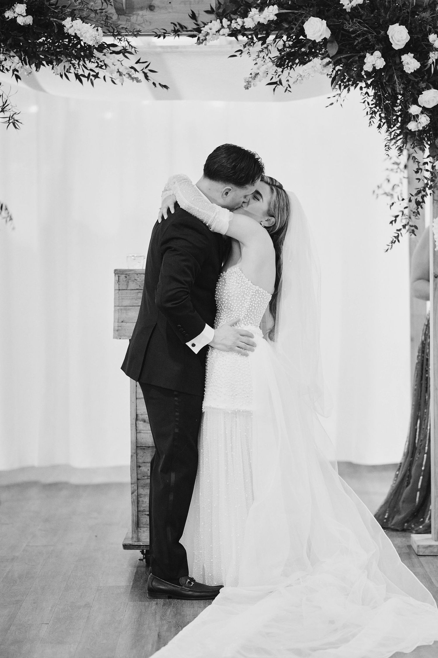 A romantic black and white photo of a couple embracing during their wedding ceremony under a floral arch.