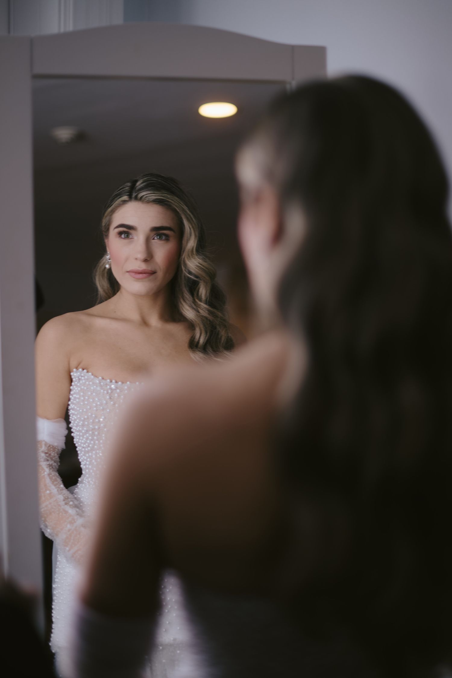 Bride in white dress with pearl details looks at her reflection through mirror on wedding day.