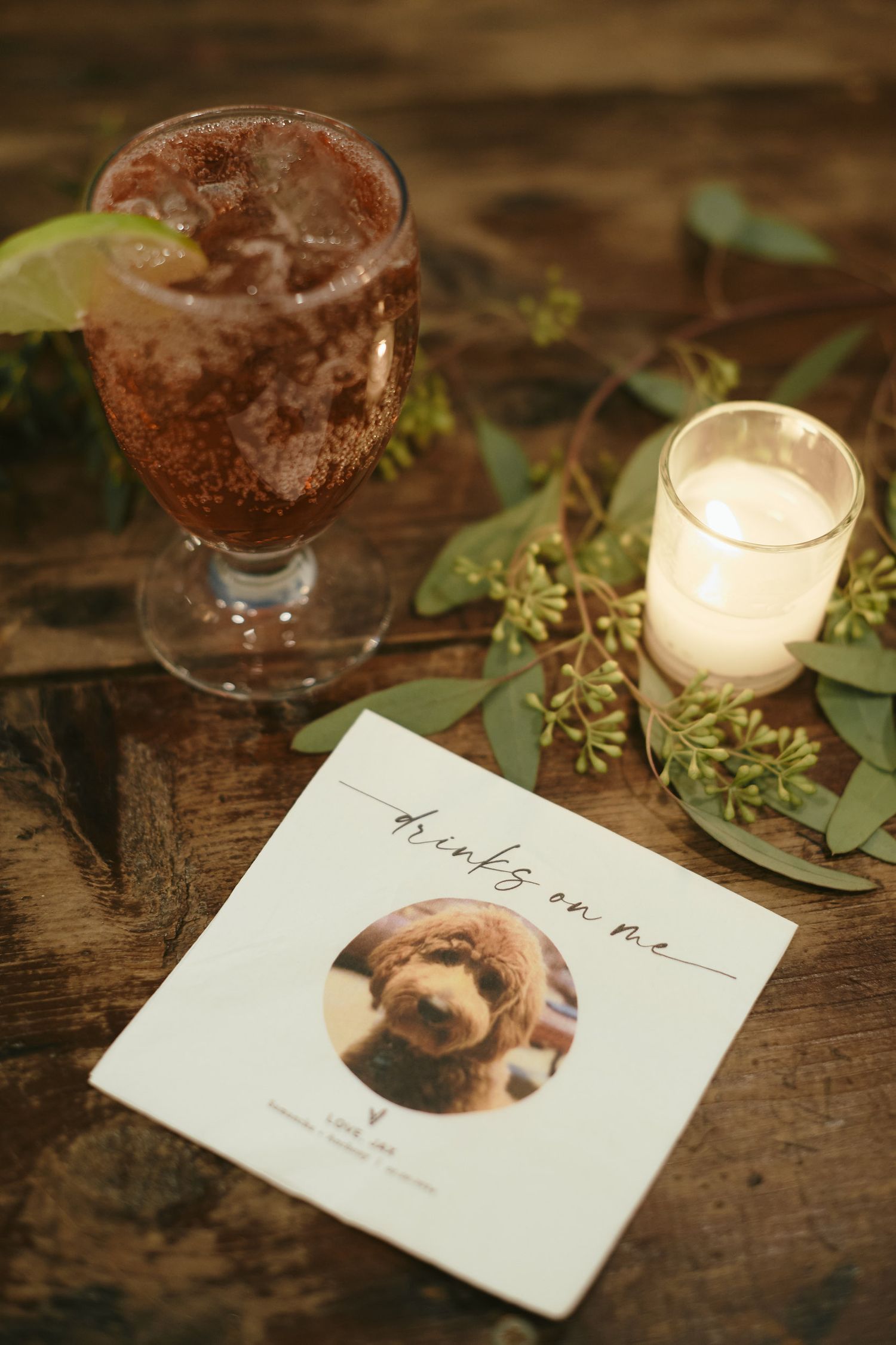 Stylish cocktail drink with lime garnish next to a white candle and printed photo card on rustic wooden table.