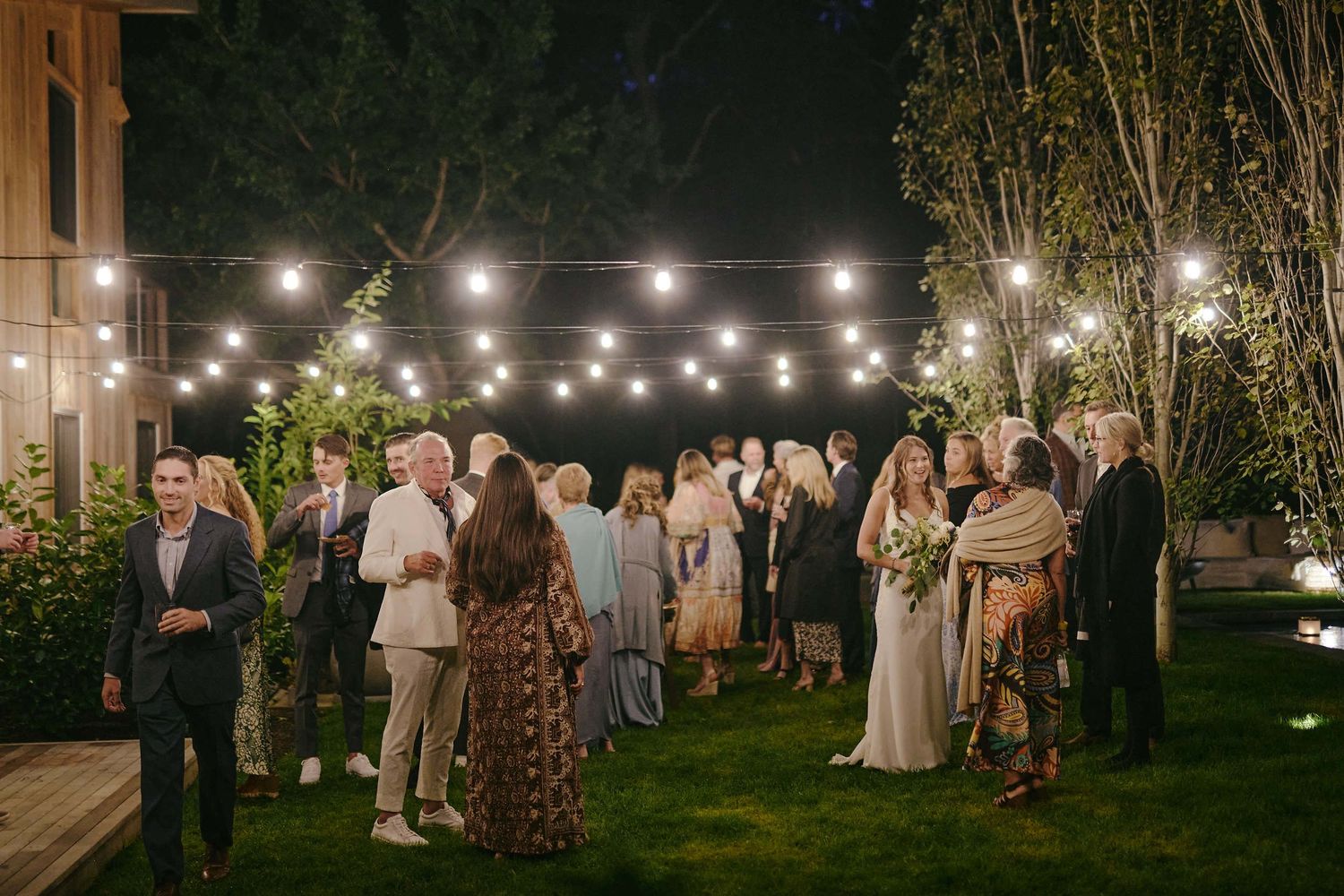 Evening garden party with guests mingling under warm string lights on a manicured lawn.