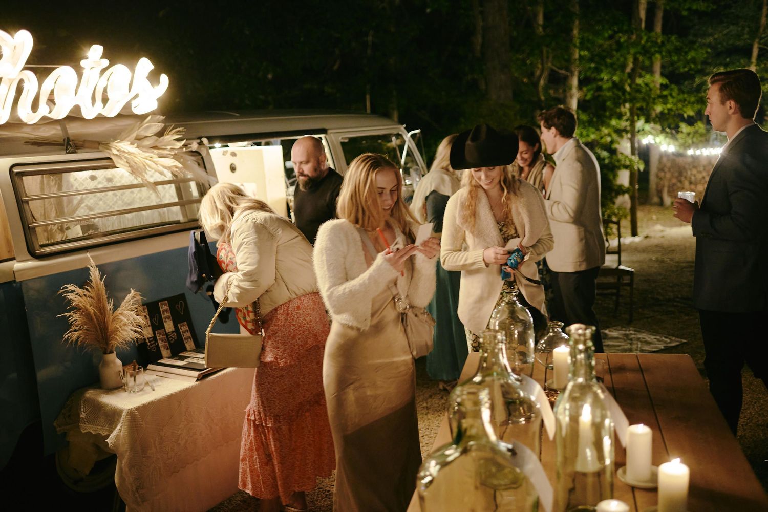Party guests gathered around a vintage camper van with string lights and wine bottles creating a festive nighttime atmosphere.