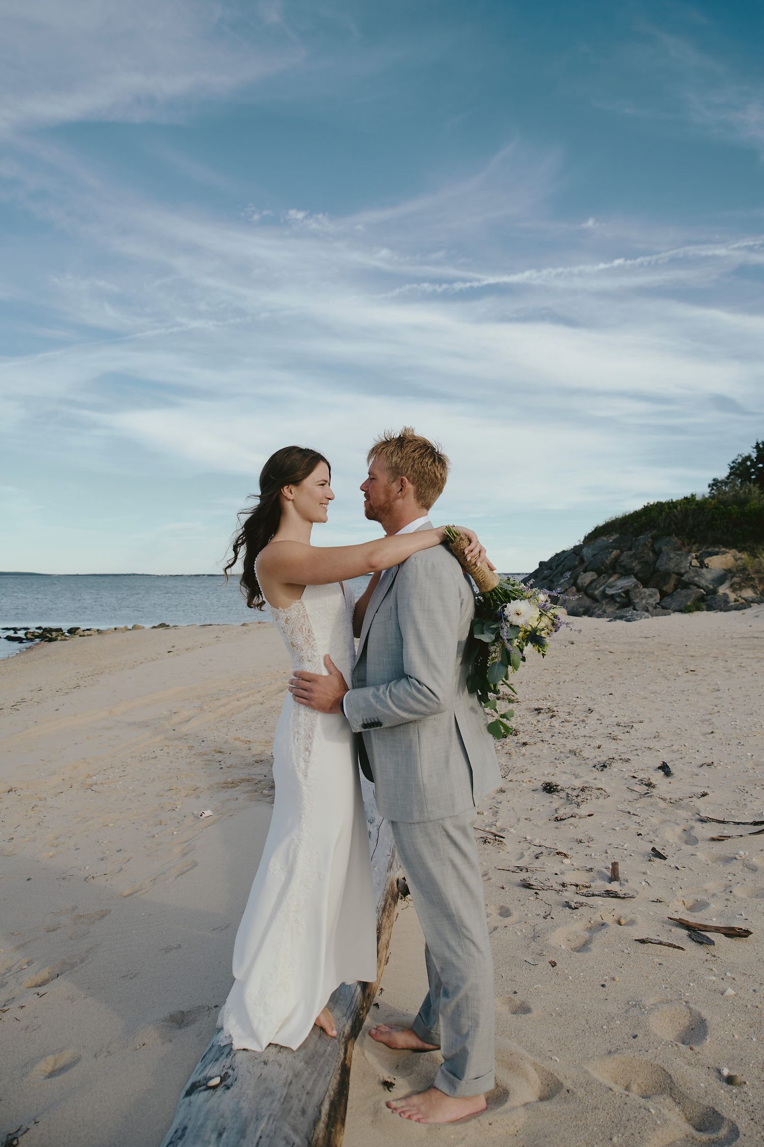 Couple embraces on sandy beach during sunset wedding ceremony with ocean and rocky cliffs in background.