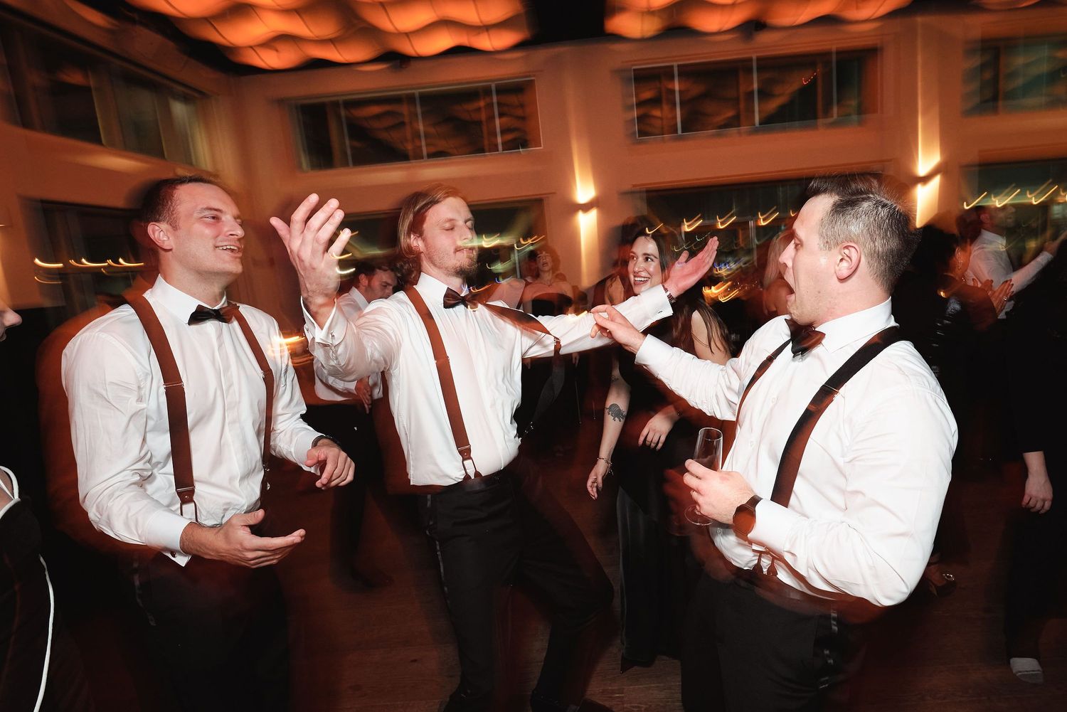Groomsmen in matching suspenders and bowties celebrate energetically on the dance floor at a wedding reception.