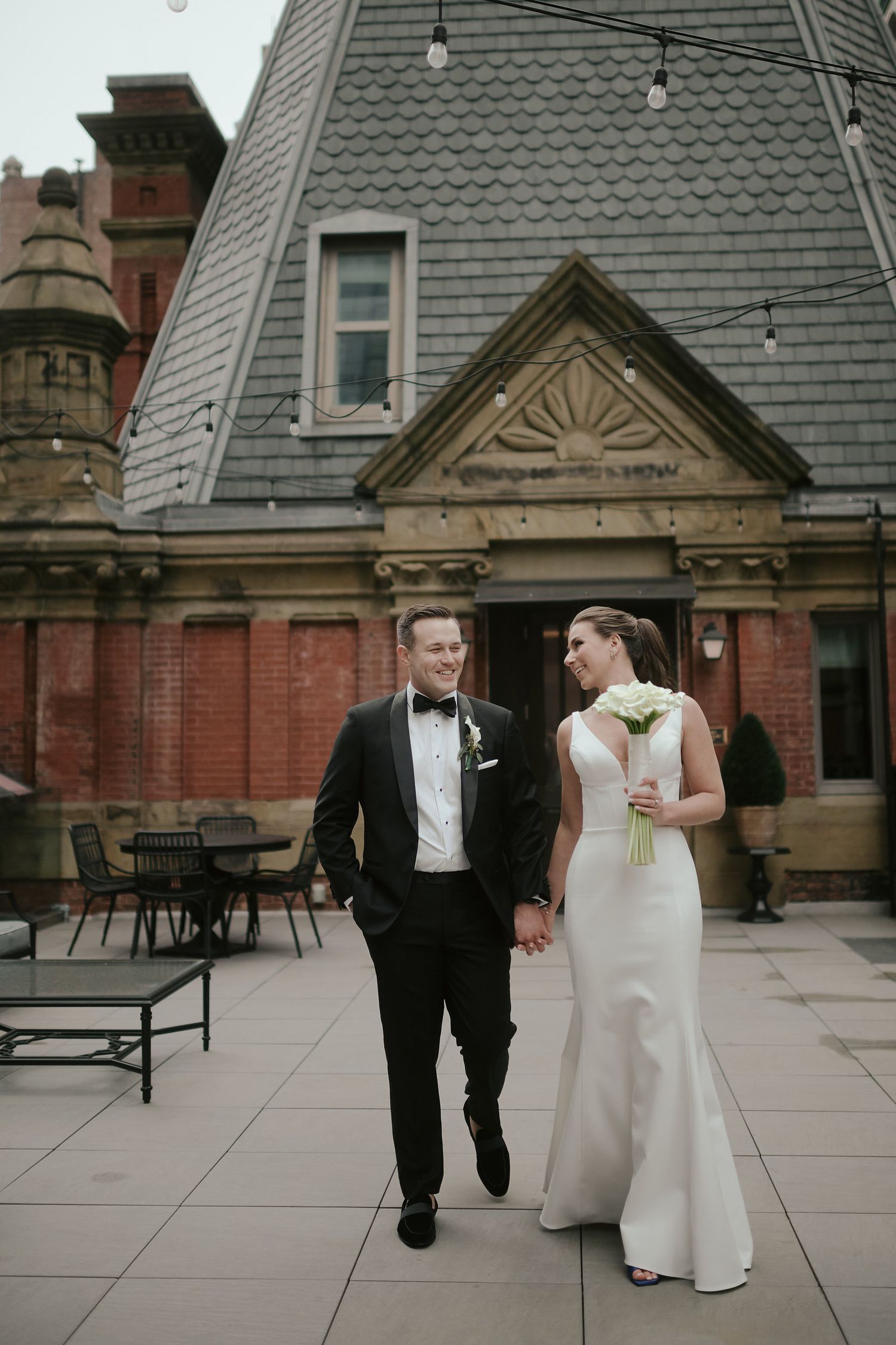 Newlywed couple walking together on a terrace with Victorian architecture in the background.