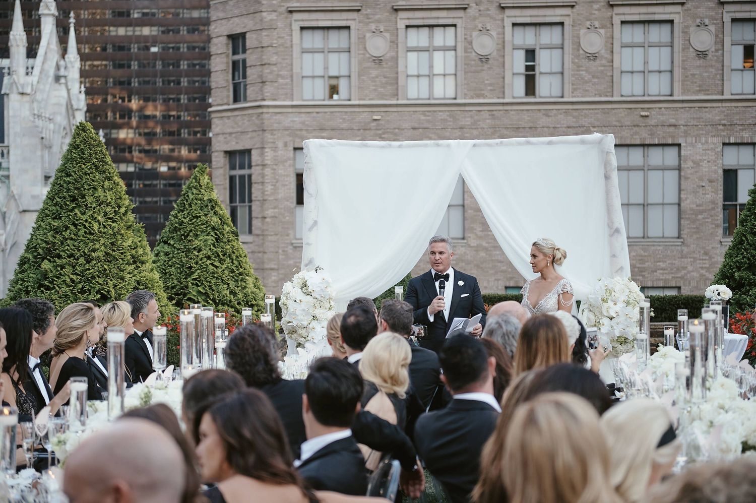 Wedding ceremony guests gathered outdoors at an elegant venue with white floral decorations.