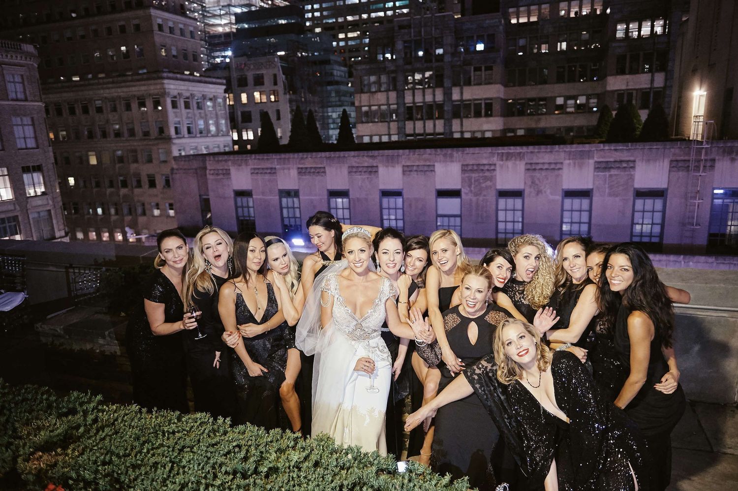 A group of formally dressed wedding guests in black attire gathered on a rooftop with city buildings in the background.