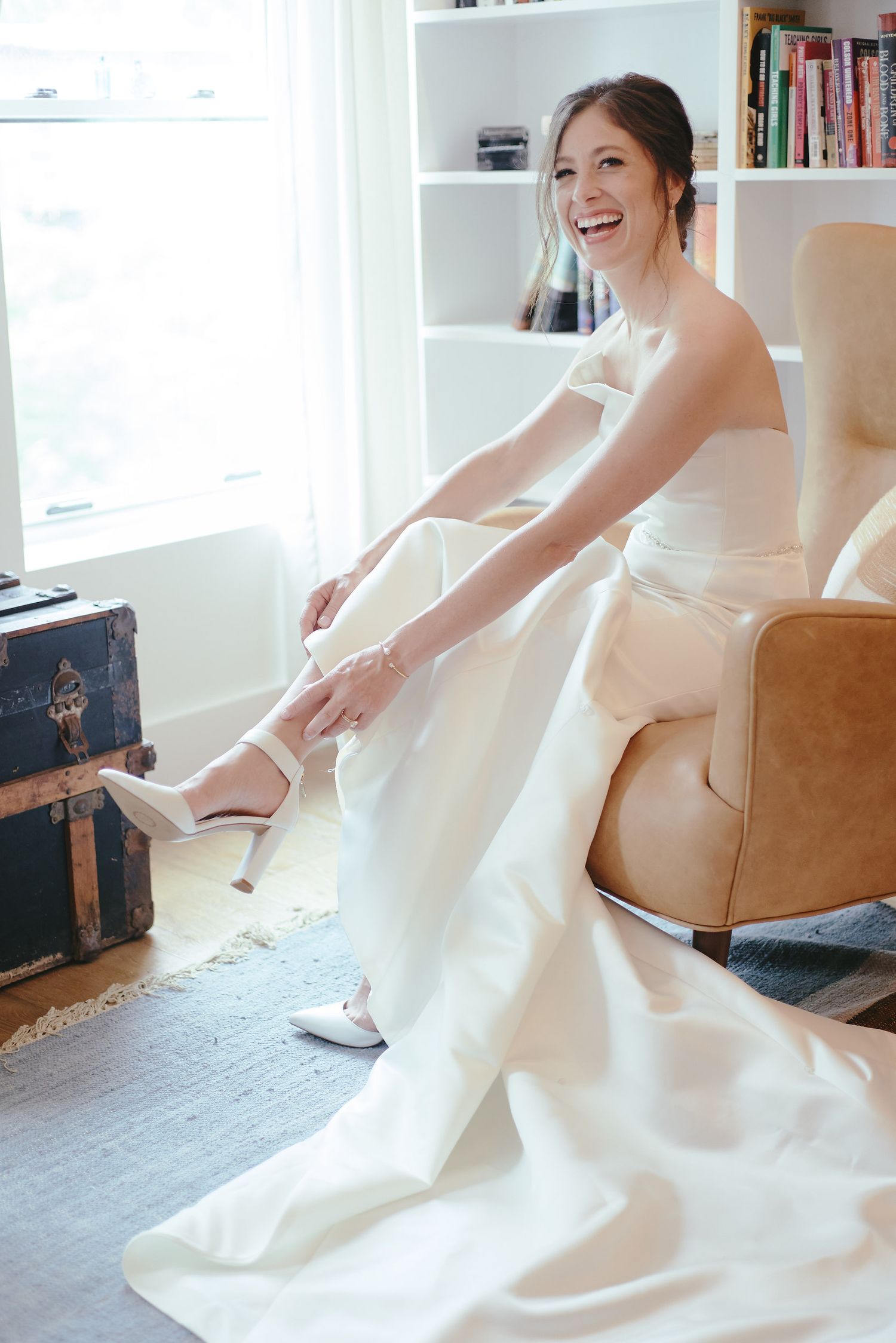 Person in white wedding dress sits on tan chair while putting on shoes.