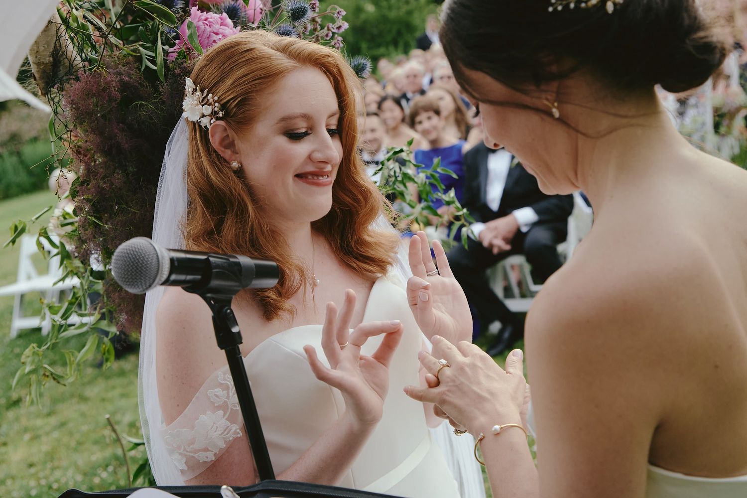 Two people share a moment during an outdoor wedding ceremony with a microphone stand and natural greenery setting.