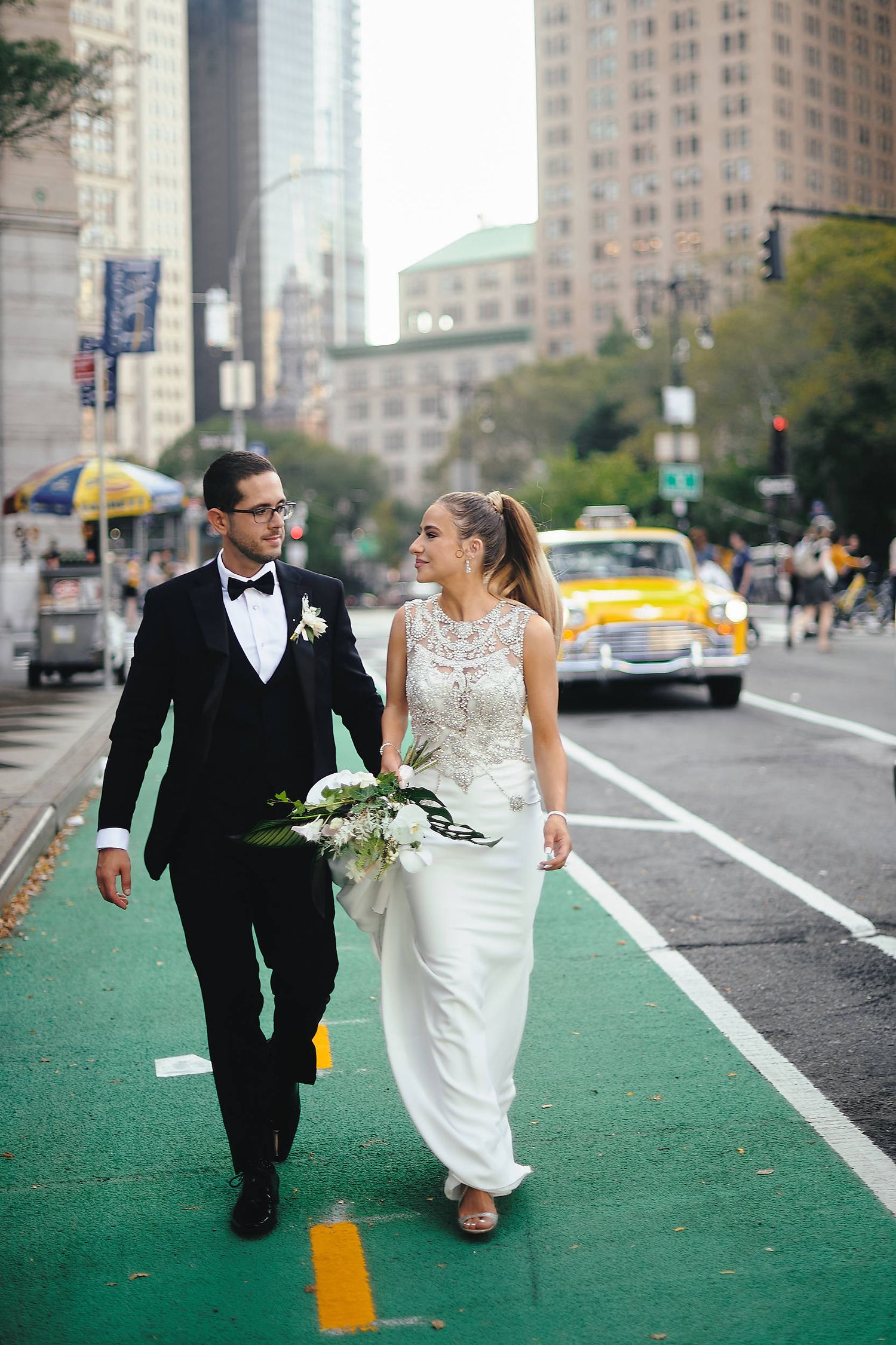 A bride and groom walk together along a green bike lane in New York City with yellow taxis and skyscrapers in background.