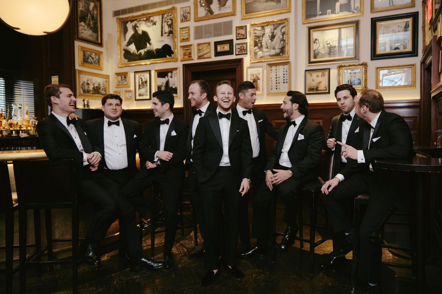 A group of groomsmen in black tuxedos gather at a dimly lit bar adorned with framed photos on the wall.