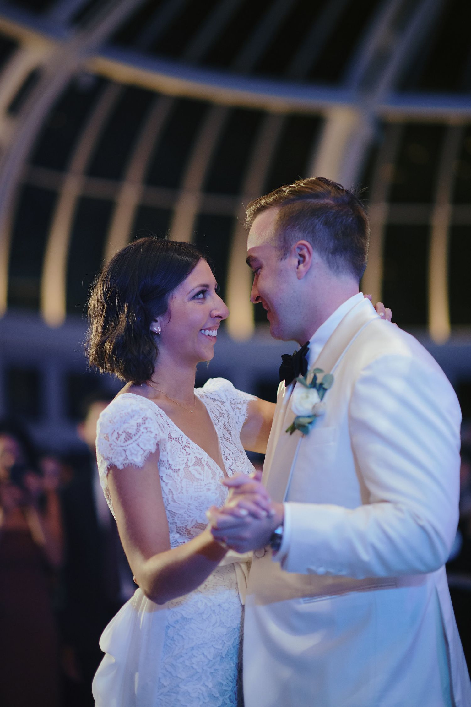 A couple shares their first dance at their wedding reception, with the bride in lace and groom in white tuxedo.
