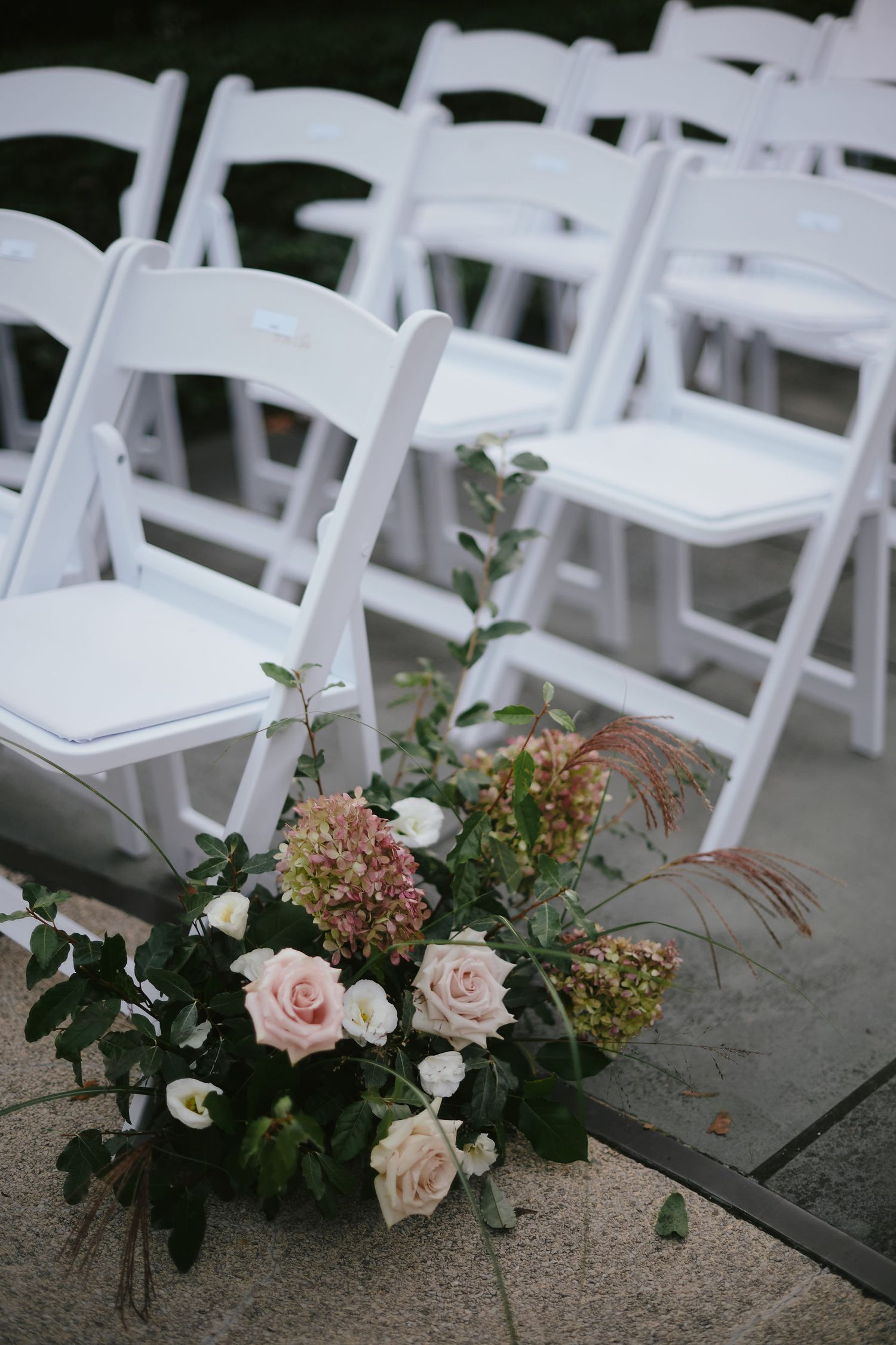 White folding chairs at a wedding ceremony with romantic blush pink roses and greenery floral arrangement on the ground.