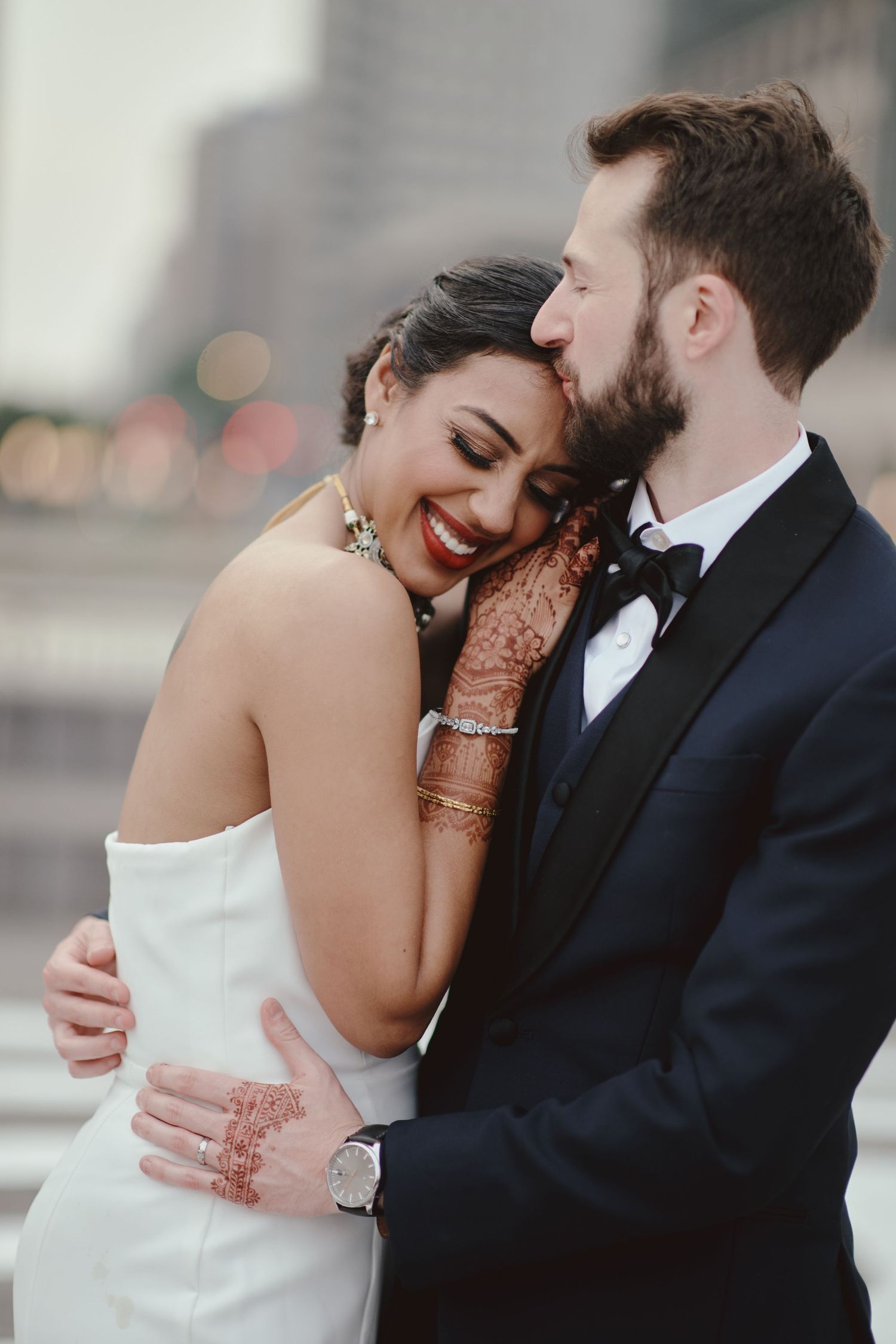 Bride with henna hands embraces groom in tuxedo, sharing romantic moment at outdoor wedding celebration.
