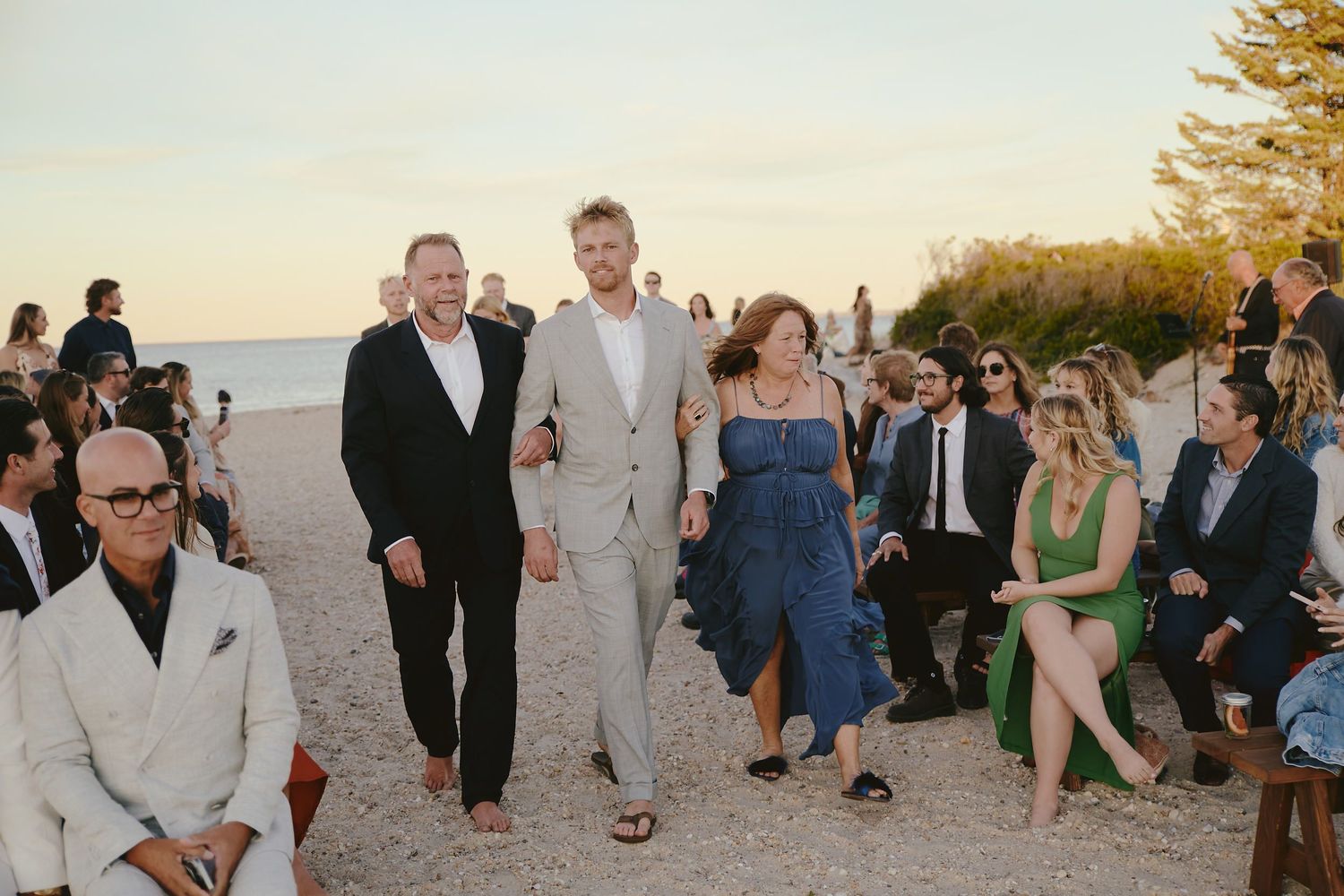 A group of wedding guests walks along an outdoor path at sunset while others are seated for a beach ceremony.