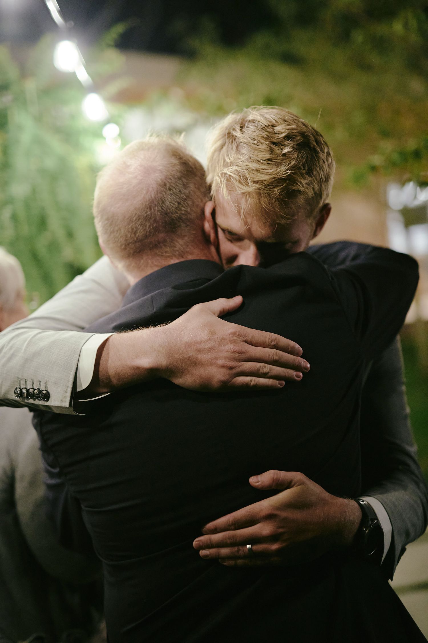 Two people share an emotional embrace at an evening event outdoors with bokeh lights in the background.