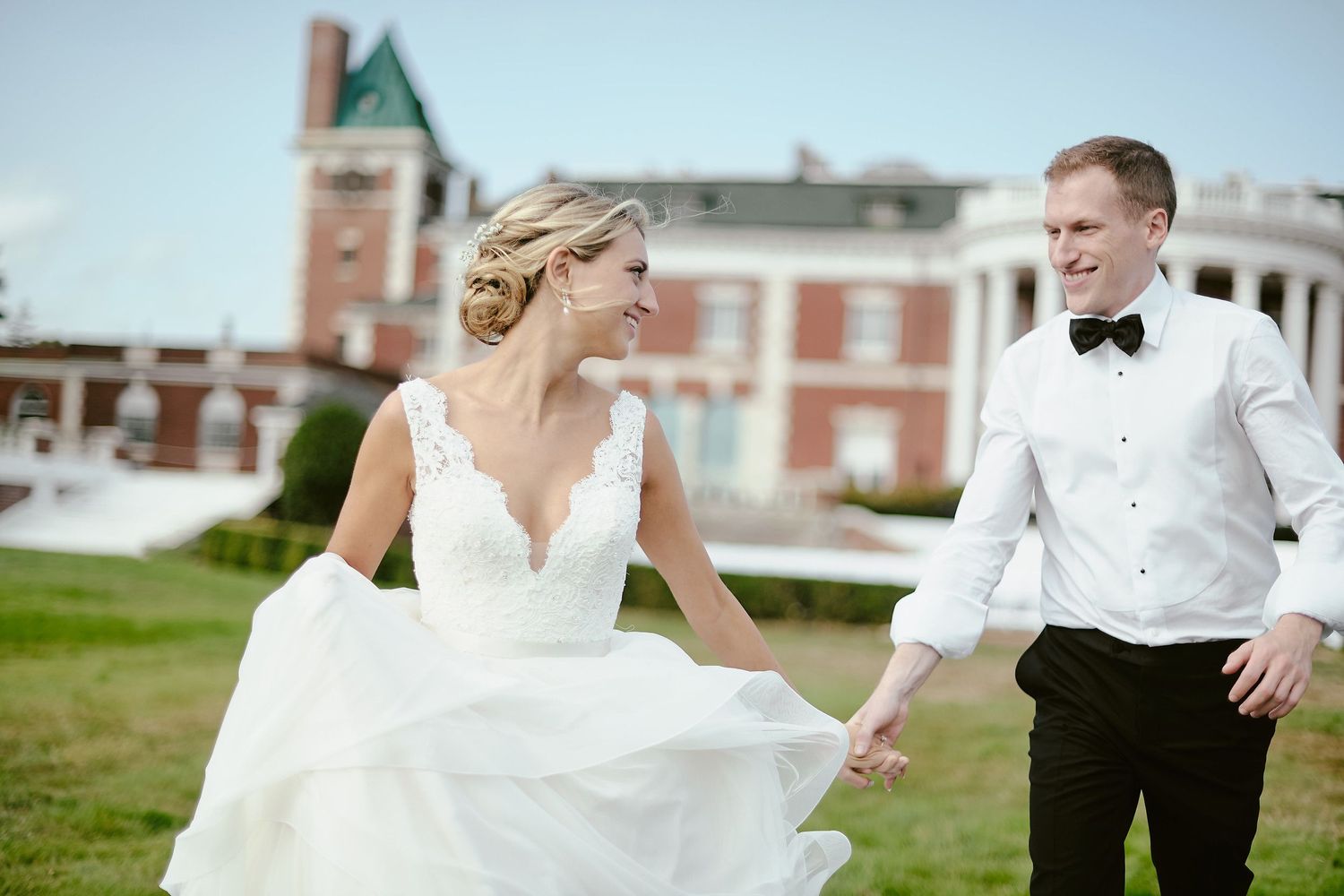 Joyful couple laughing together on lawn with historic mansion in background during wedding photoshoot.