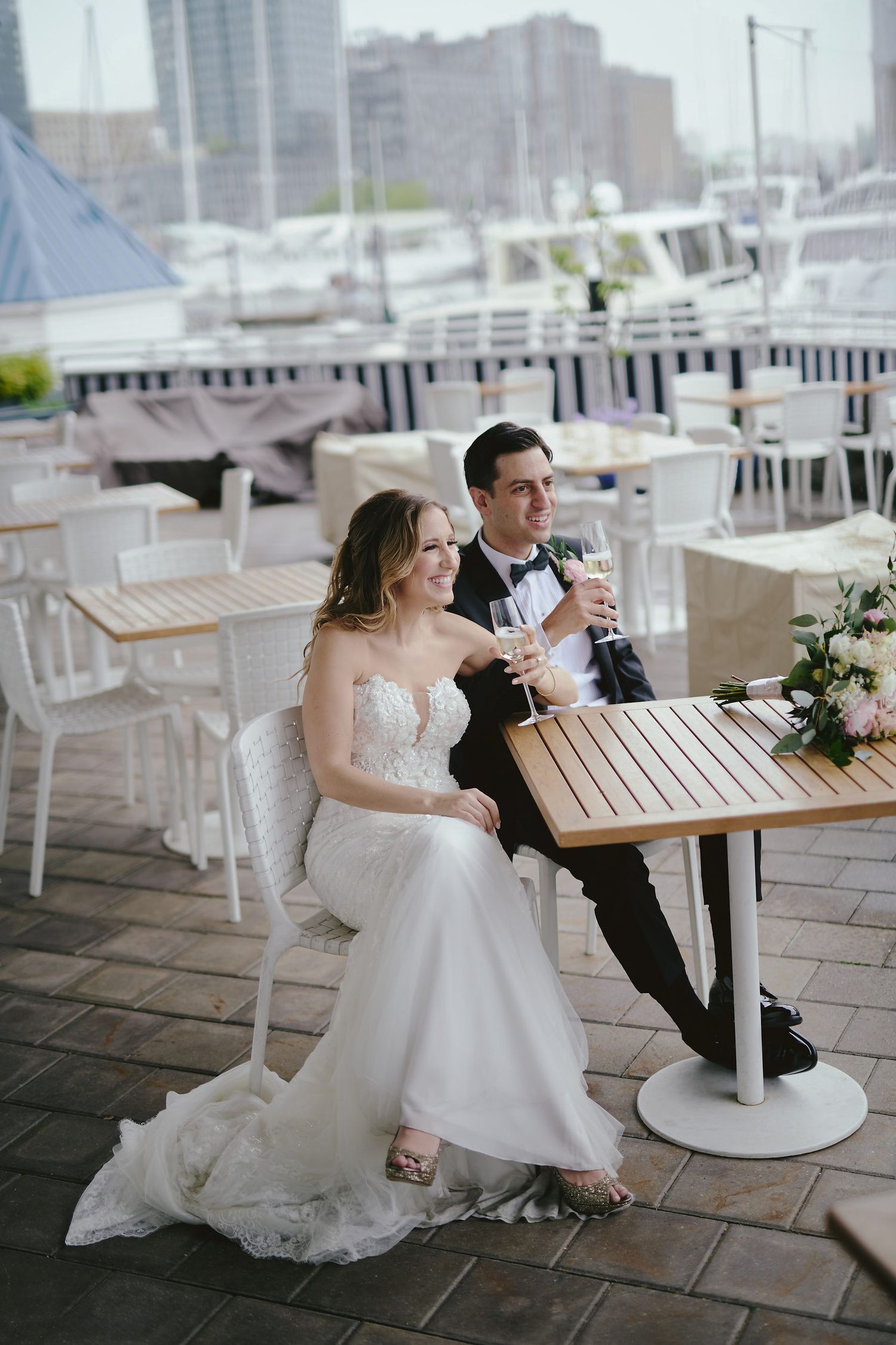 A couple in wedding attire shares drinks at an outdoor waterfront restaurant patio with boats in the background.