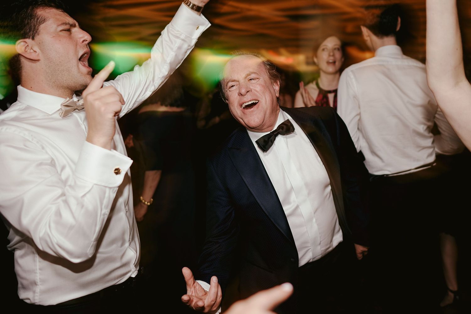 Party guests celebrate exuberantly on a dimly lit dance floor wearing formal black and white attire.