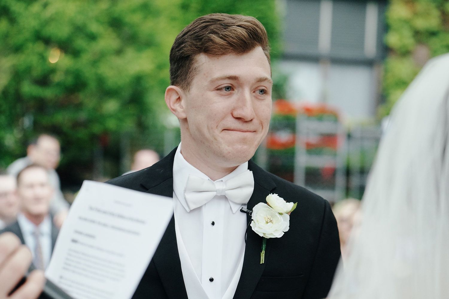 A groom in a black tuxedo with white boutonniere smiles during an outdoor wedding ceremony.