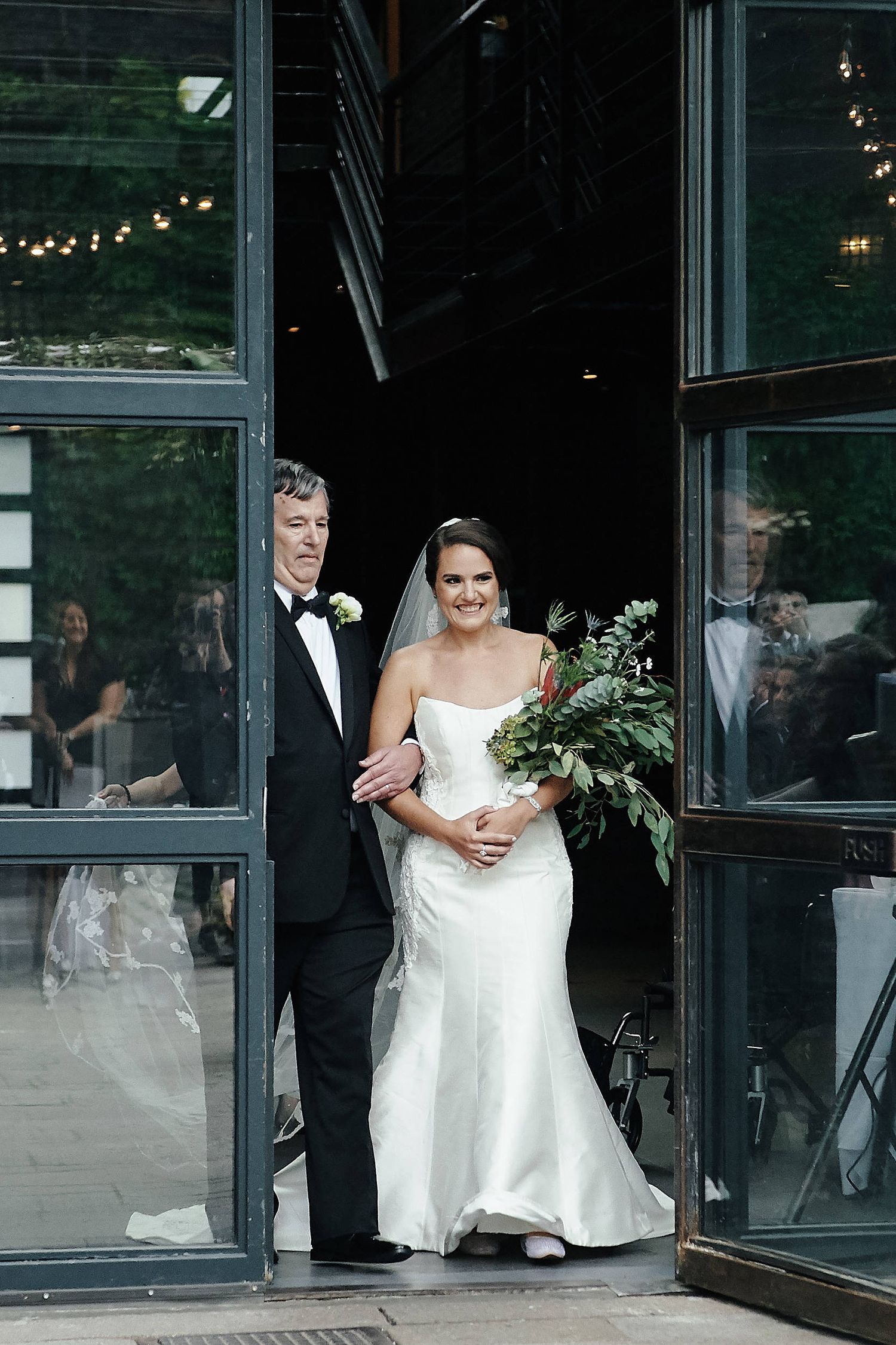 A couple emerges from modern glass doors at their wedding venue with bride holding an elegant greenery bouquet.