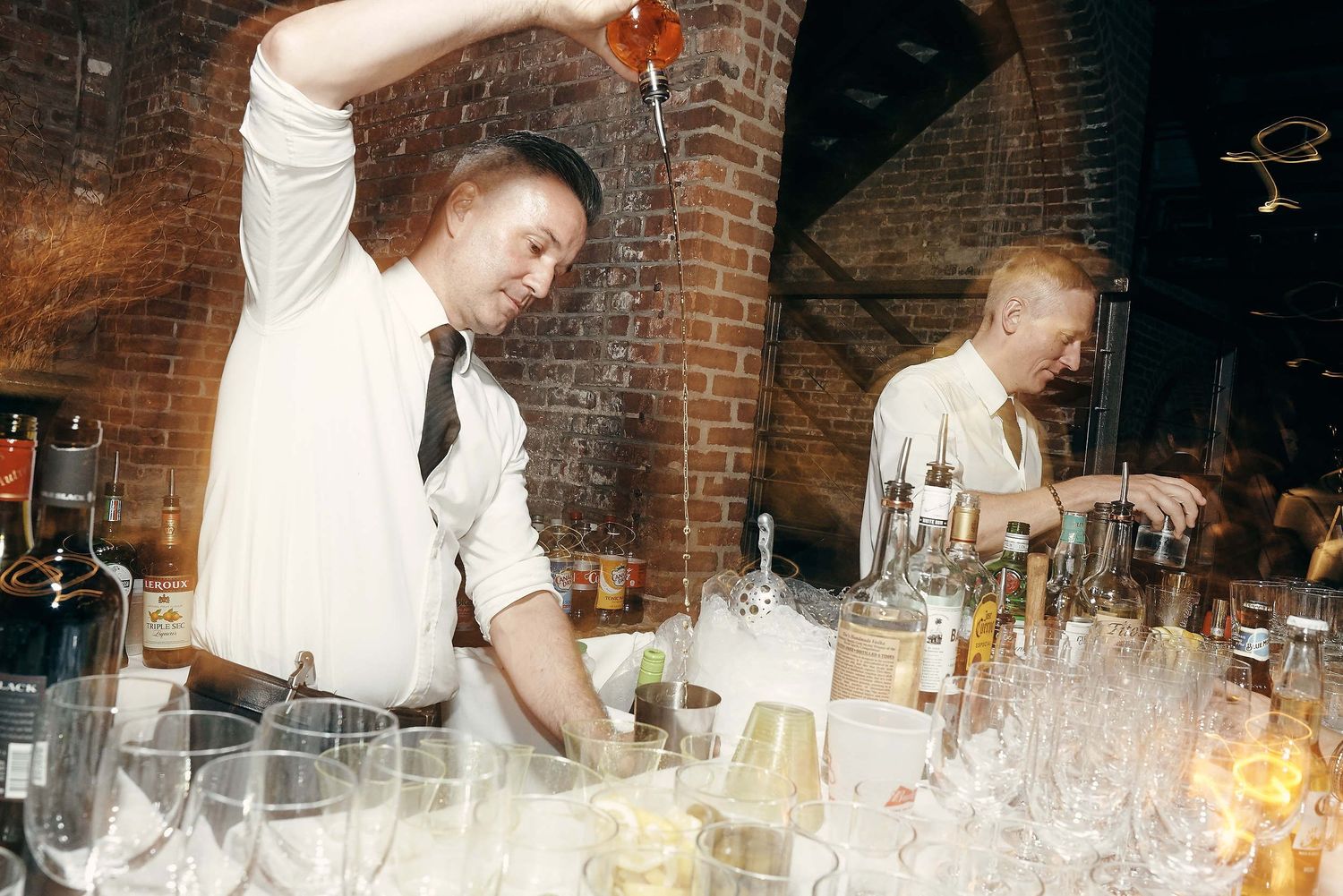 Bartenders pouring cocktails and mixing drinks behind bar at event with brick wall backdrop.