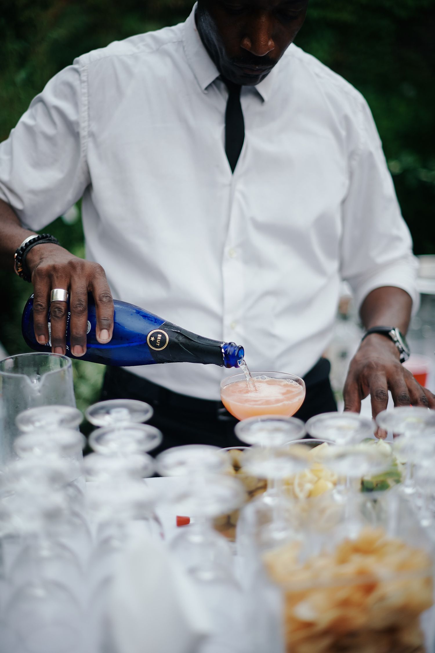 Professional bartender in white dress shirt and black tie pours a pink cocktail at an elegant event service station.