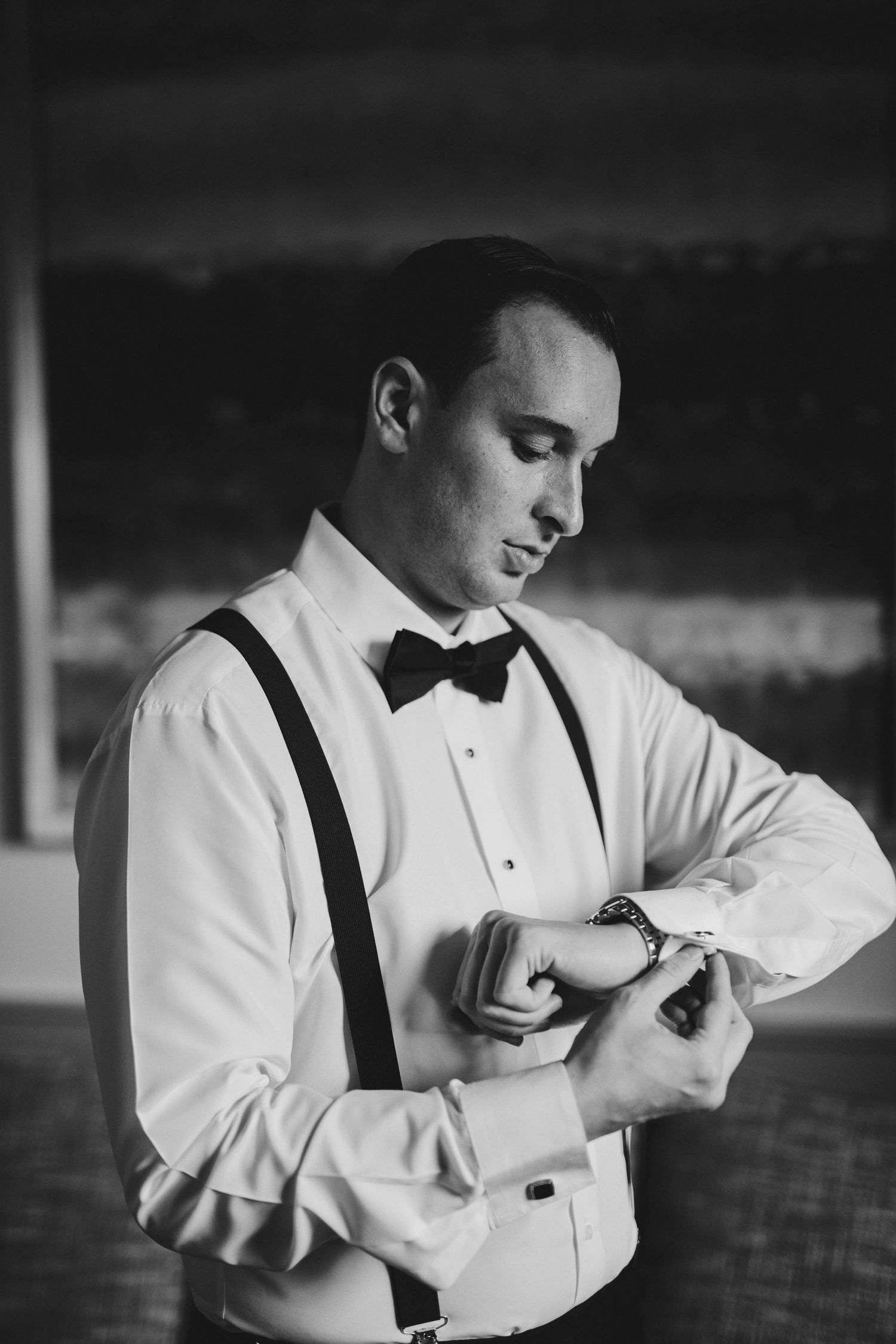 Groom in formal attire with bowtie and suspenders checks his watch in elegant black and white photograph.