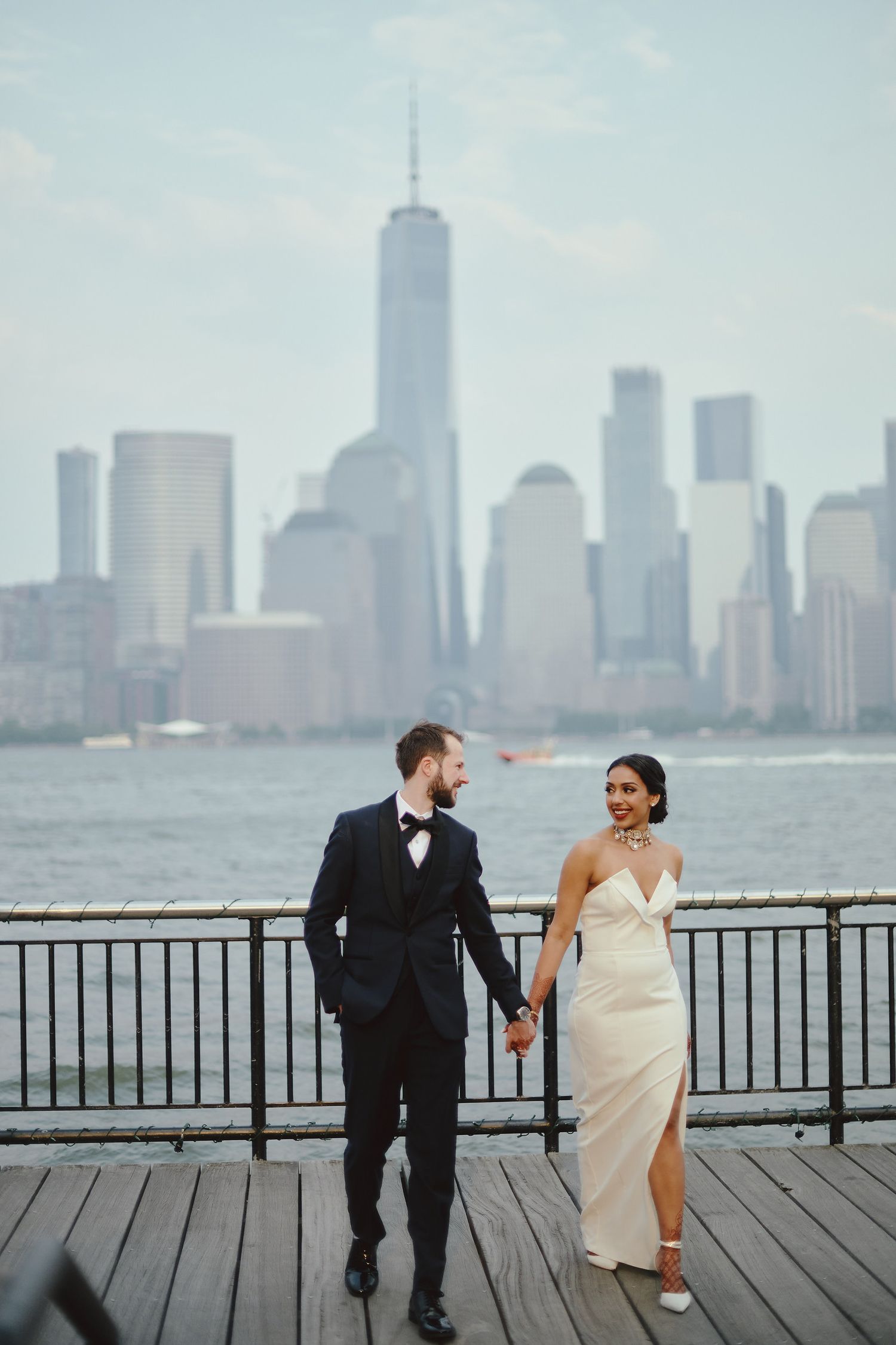 A couple in wedding attire holds hands while walking on a pier with the New York City skyline in the background.