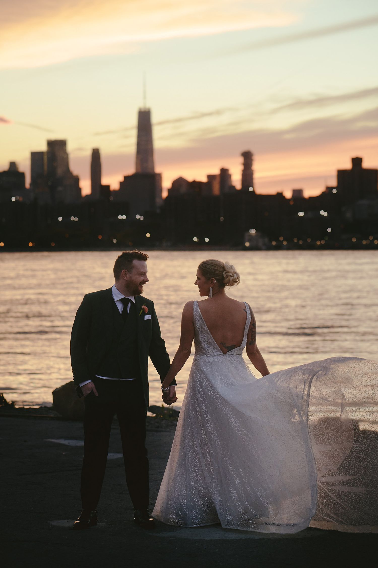 A couple on a pier viewing the New York skyline silhouette during golden sunset, white wedding dress flowing in breeze.