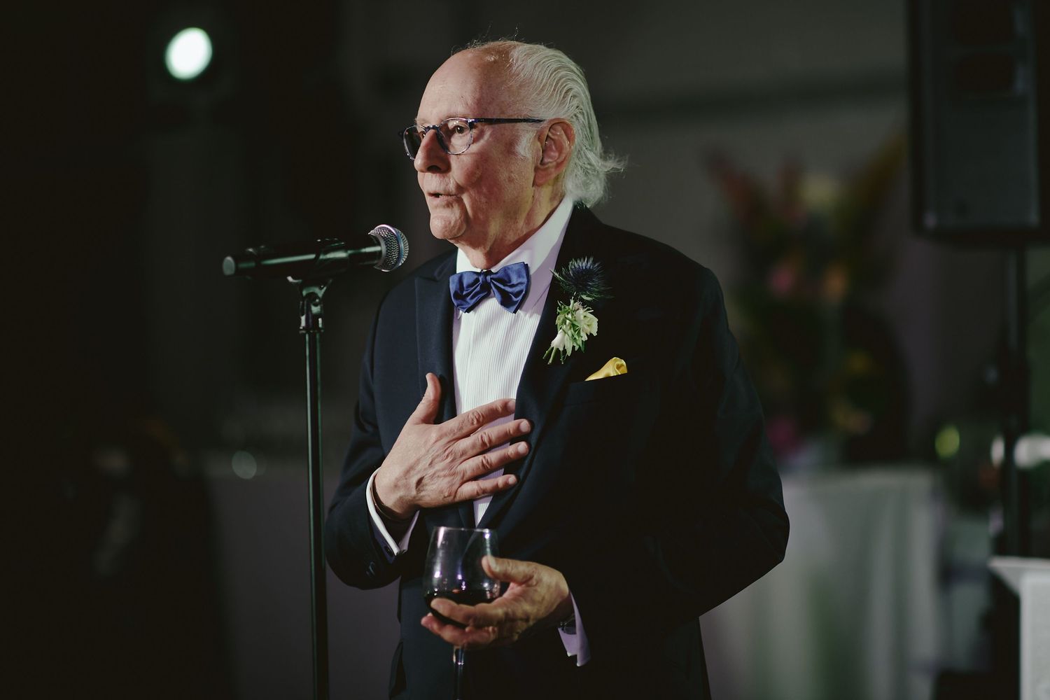 An elegant senior gentleman in a tuxedo and bow tie speaks emotionally into a microphone at an evening event.