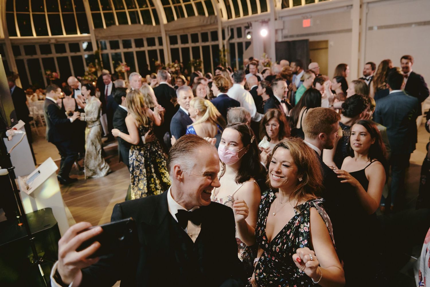 A lively crowd of party guests dance and celebrate at an elegant indoor reception with warm lighting.