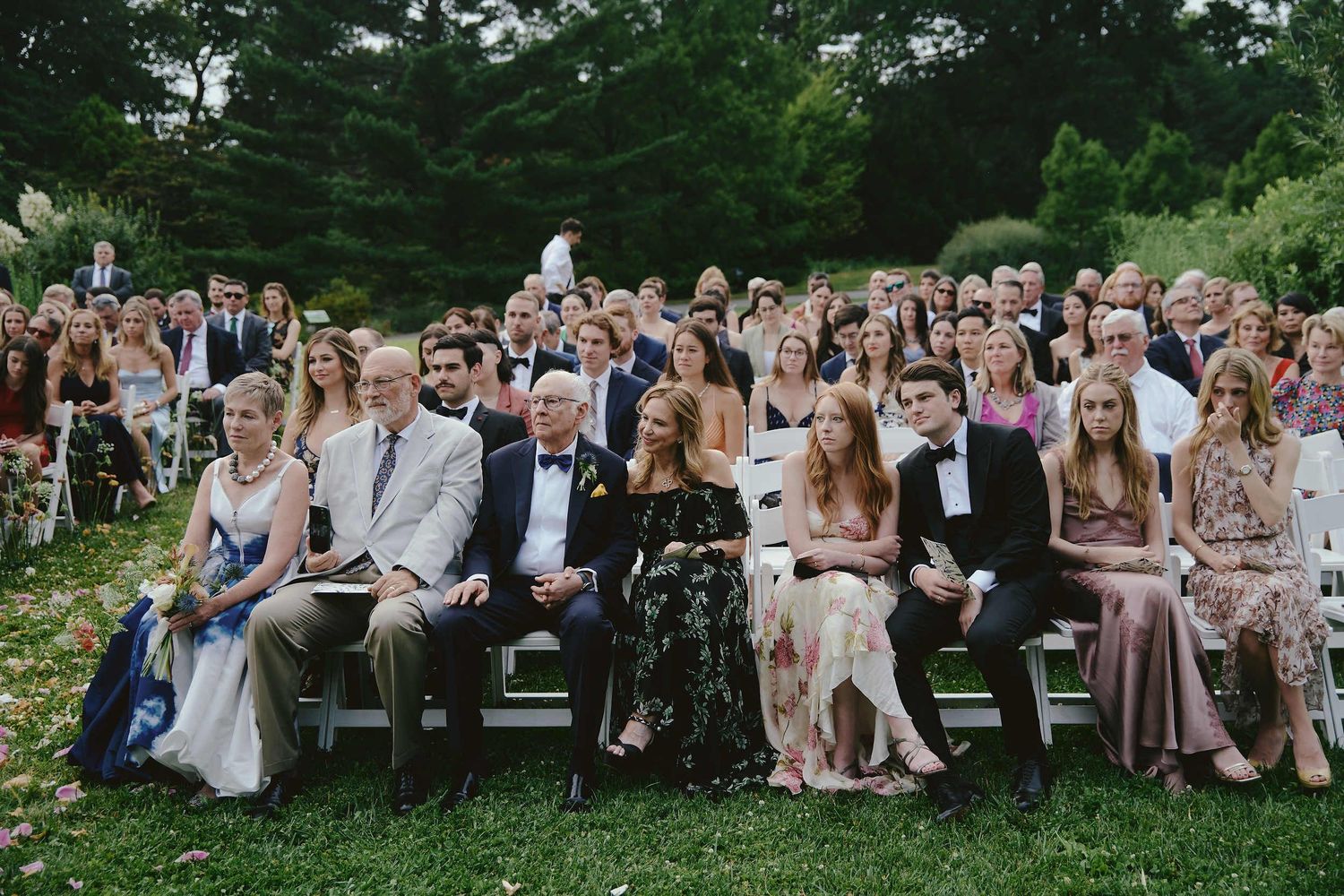 Wedding guests seated in rows of white chairs on a green lawn for an outdoor summer ceremony.