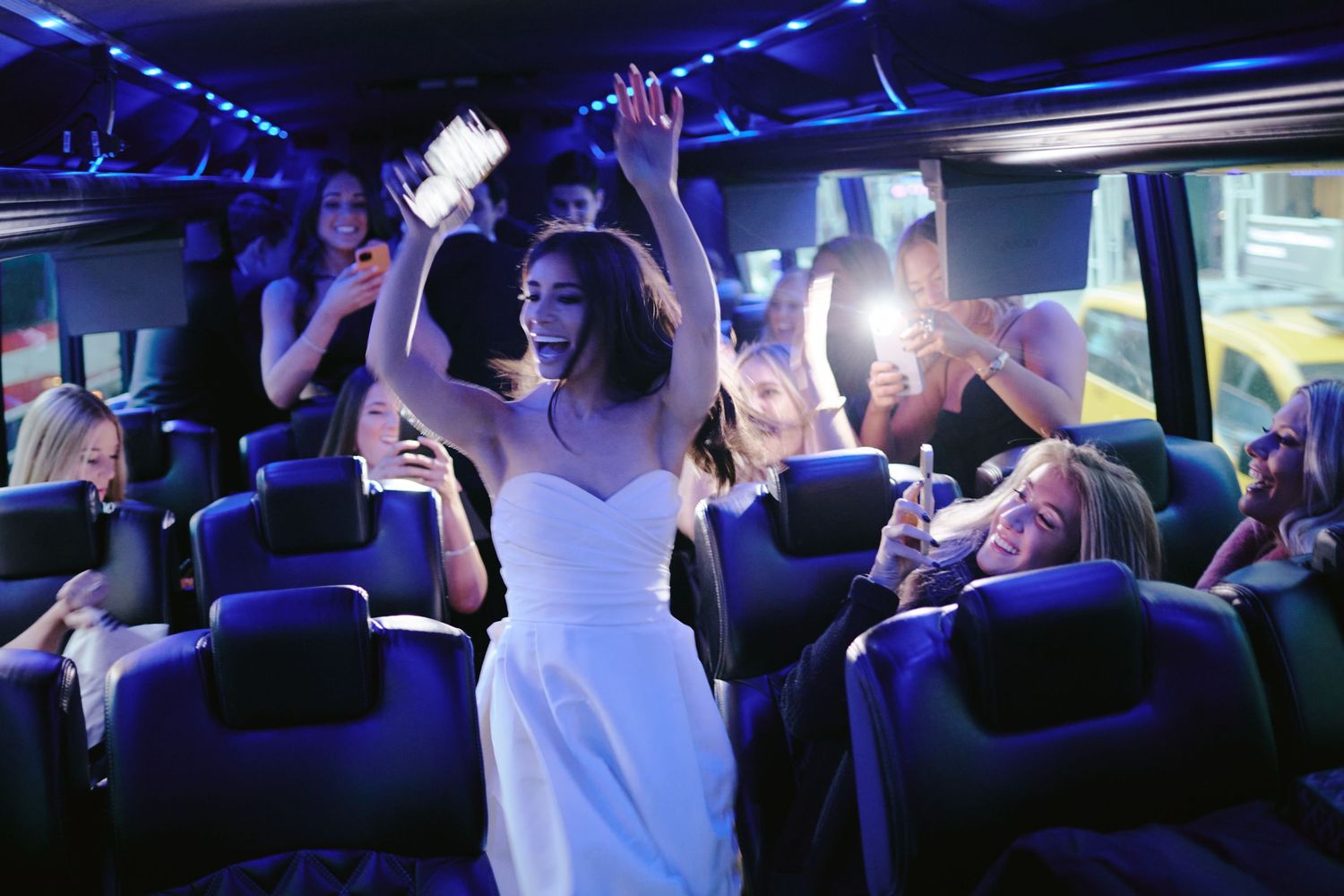 People dancing and celebrating inside a party bus with colorful blue lighting and plush seating.