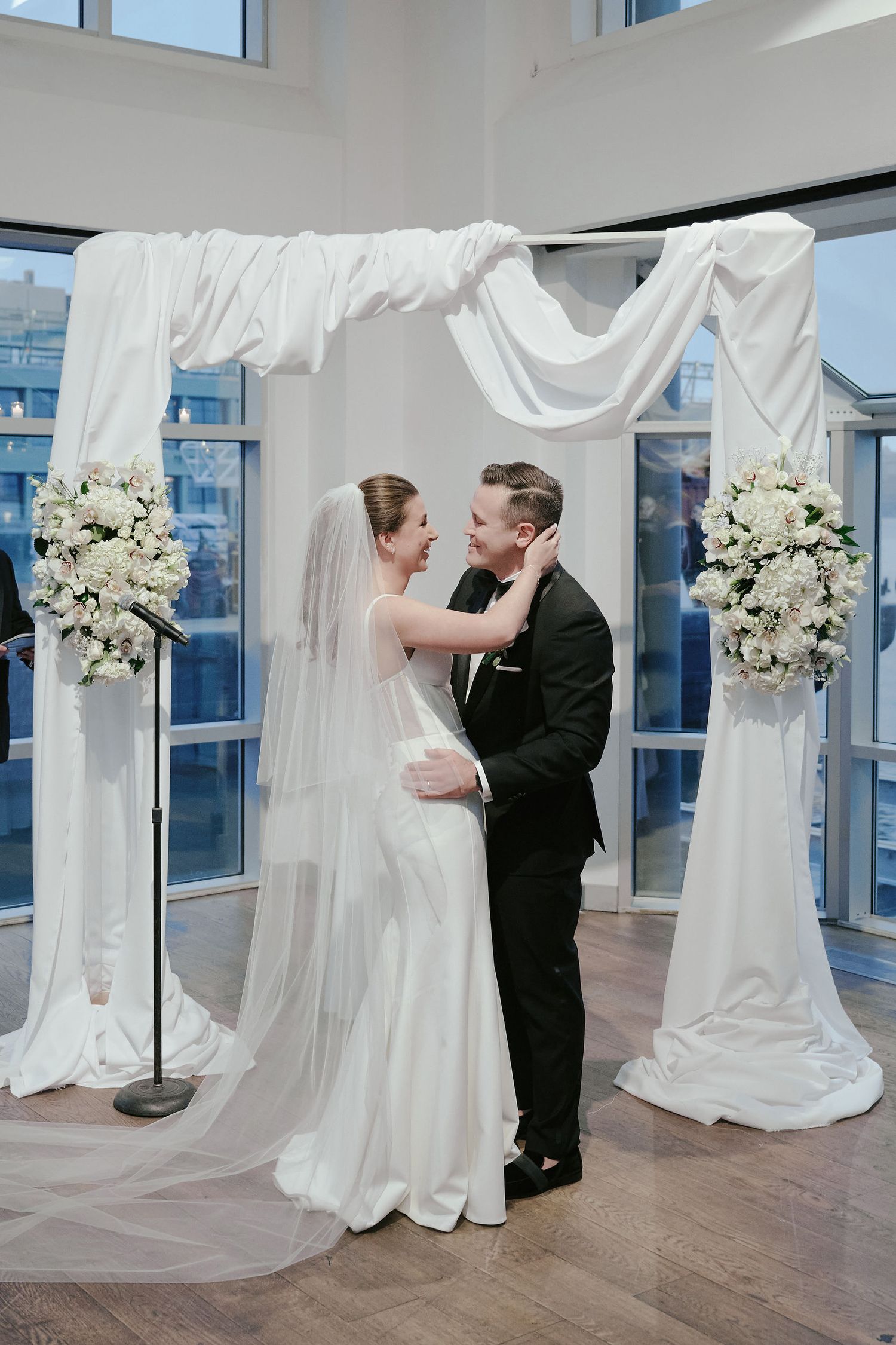 Elegant wedding ceremony under white draped fabric arch with floral arrangements in bright sunlit room with hardwood floors.