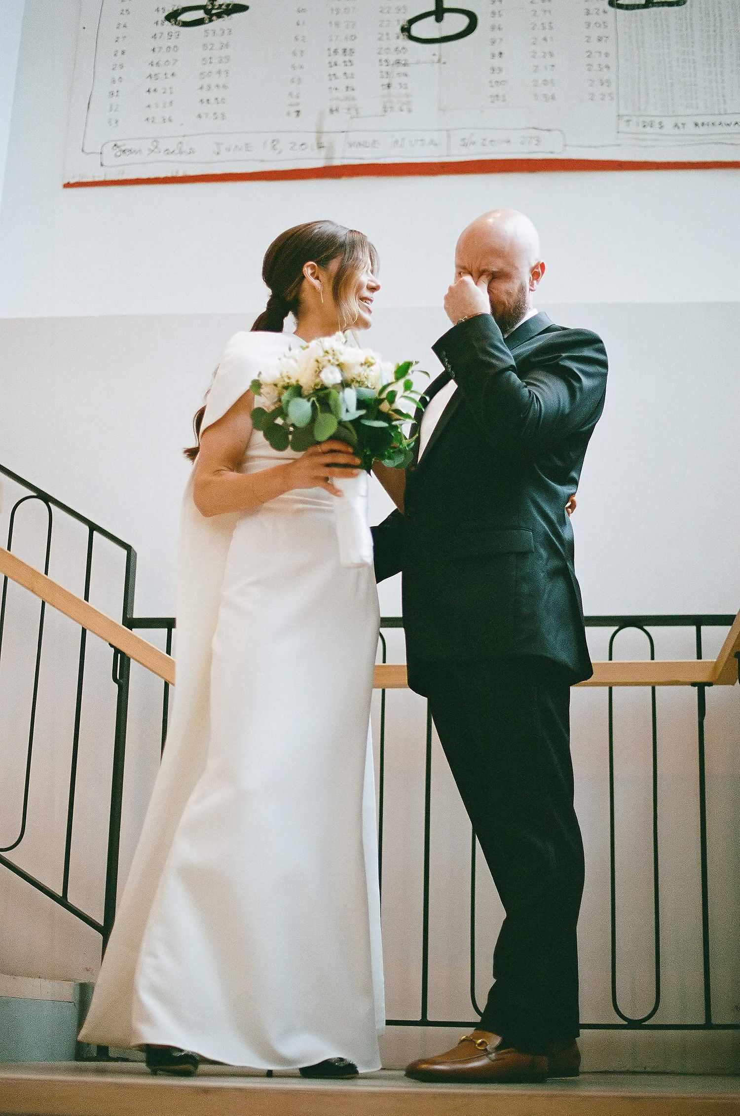 A touching wedding moment on a staircase as a couple in formal attire shares an emotional interaction with a white bouquet.