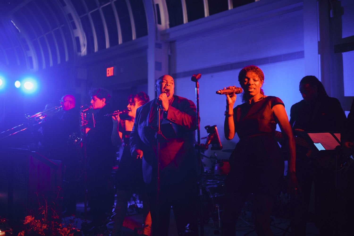 Concert performers sing on stage in moody blue and red lighting during a live music performance.