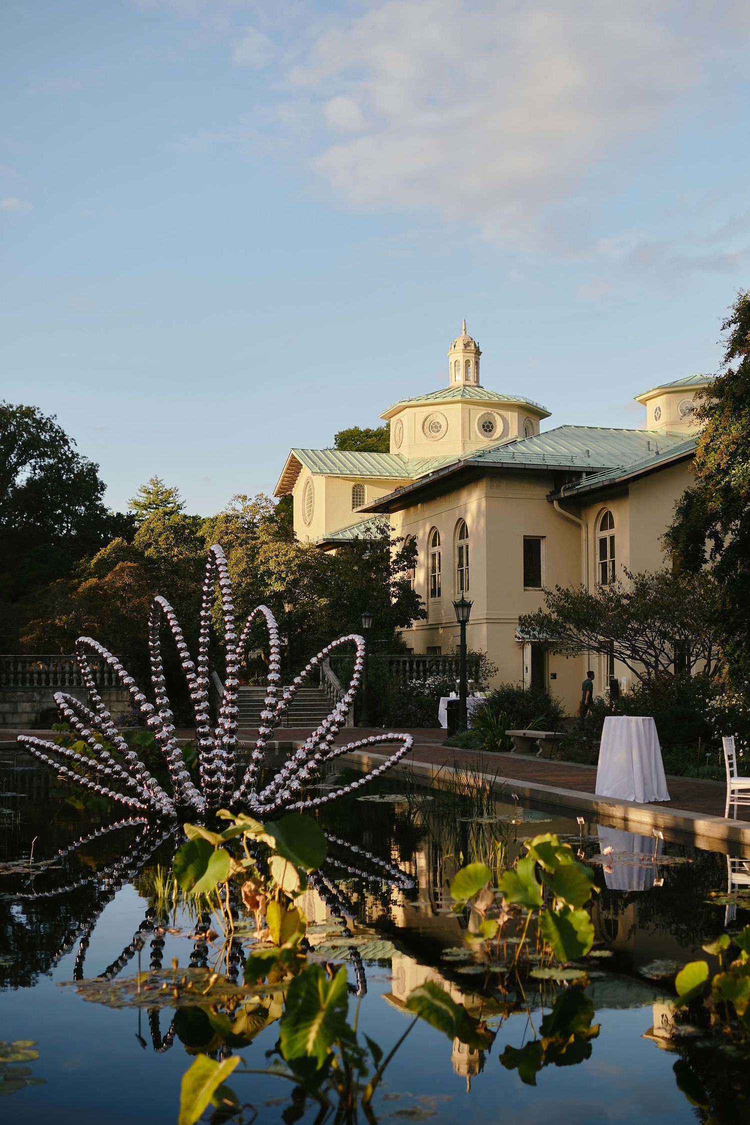 Illuminated decorative metal lotus sculpture reflects in pool water outside elegant villa building at sunset.