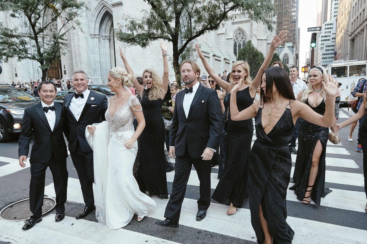 Wedding party celebrates joyfully on city street with group members in formal black and white attire.