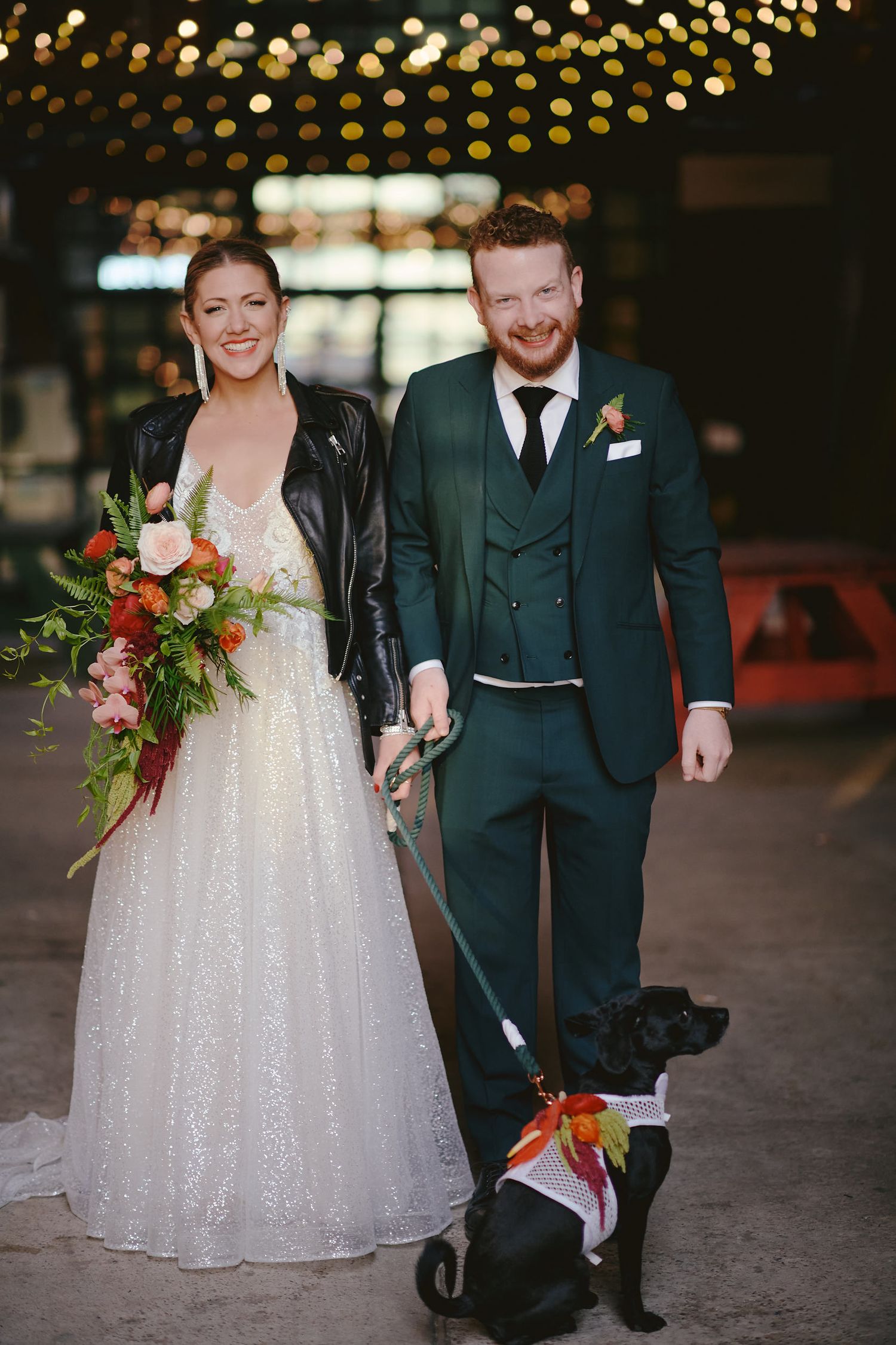 A couple in wedding attire stands together with their dog under string lights at an urban venue.