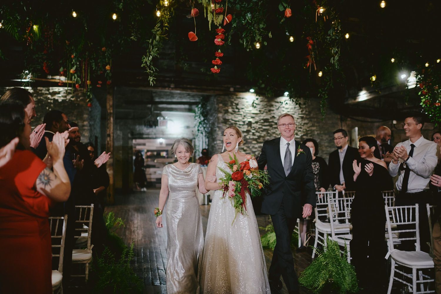Wedding processional at night under twinkling string lights and greenery in an outdoor venue setting.
