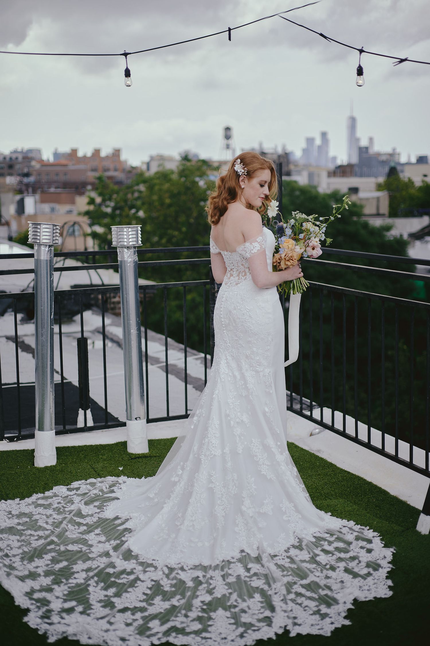 A bride in a lace wedding gown with long train stands on a rooftop terrace with city skyline view at dusk.