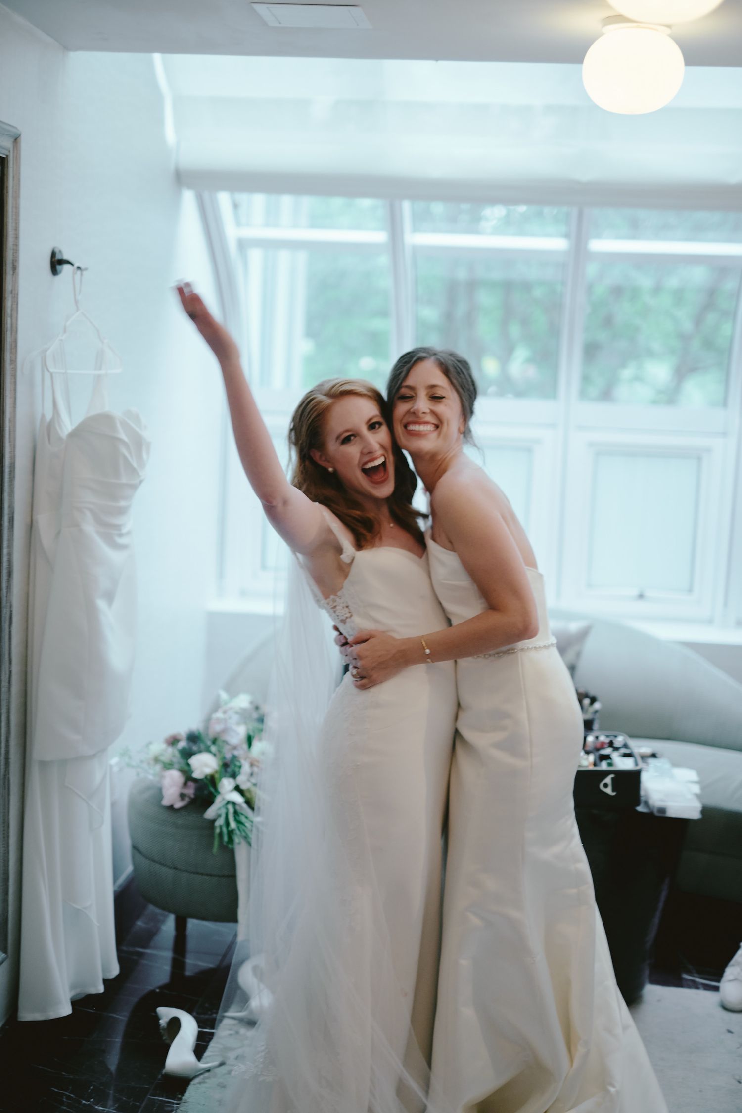 Two people in elegant white wedding dresses share a joyful embrace in a bright room with a window backdrop.