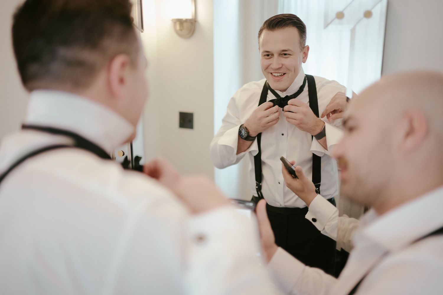 Three groomsmen in white shirts and suspenders help each other get ready before a wedding ceremony.