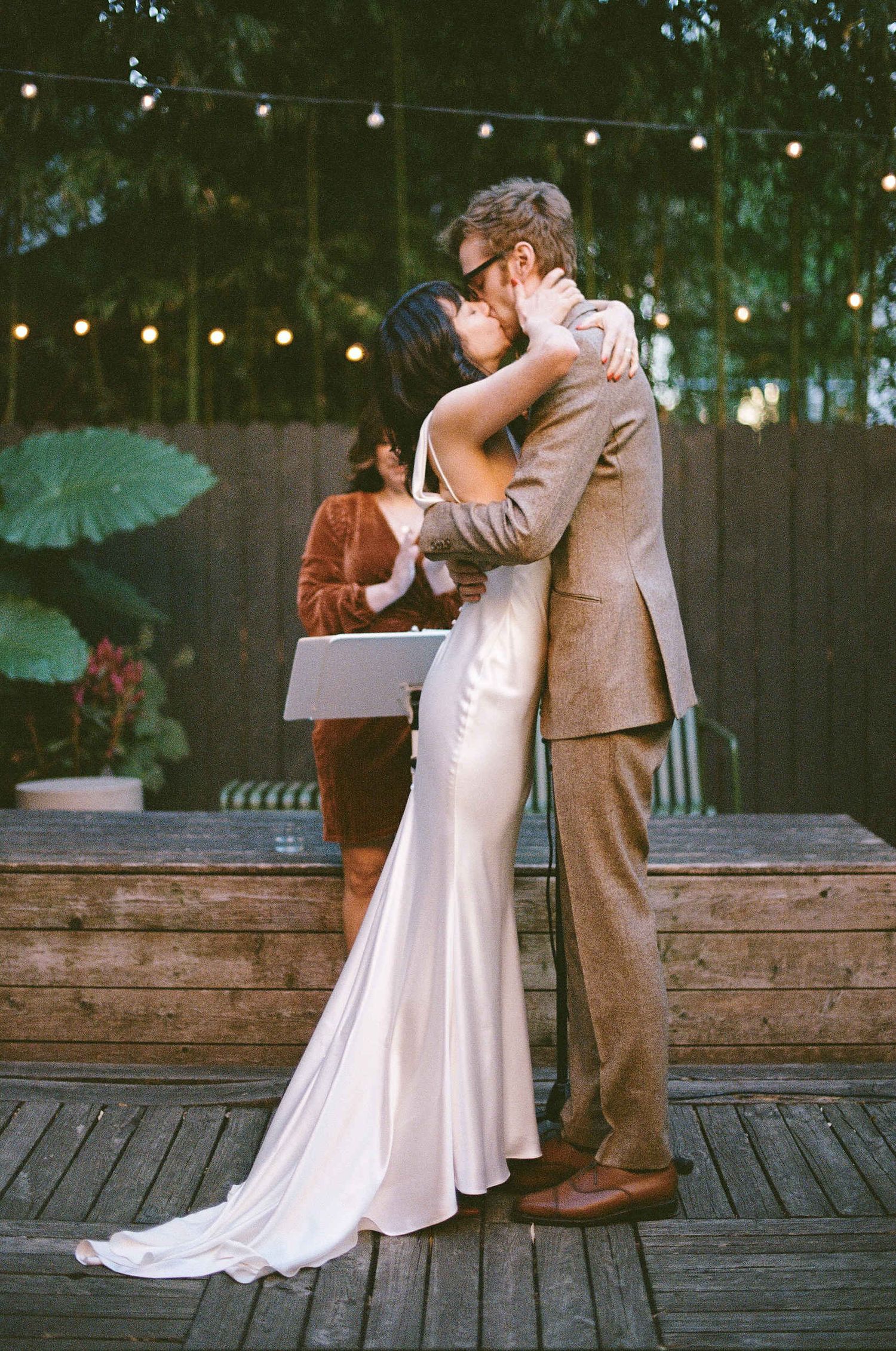 A couple embraces during their wedding ceremony on a wooden deck with string lights and parasols in the background.