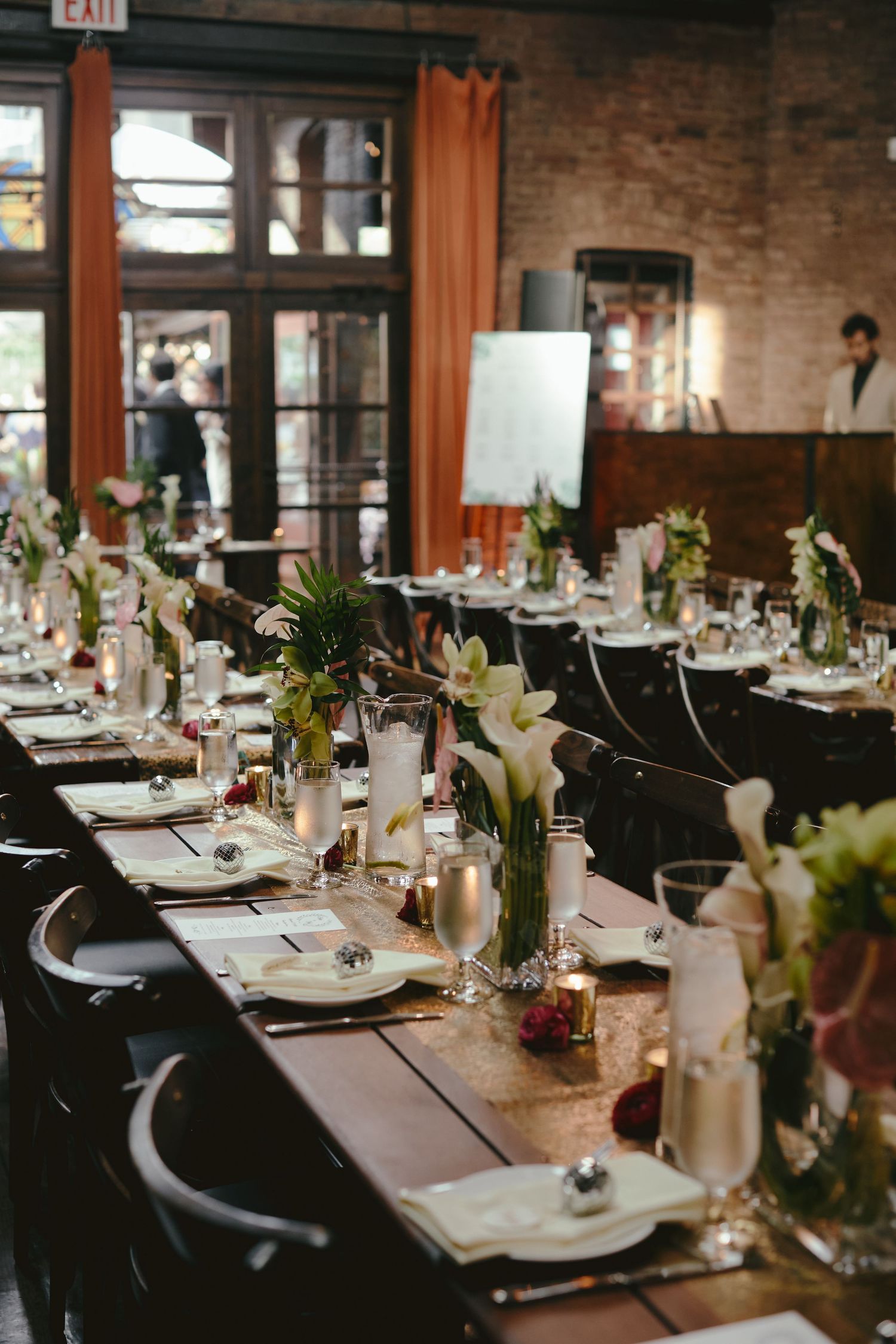 An elegant rustic wedding reception table set with white lilies, glassware and candles in an industrial brick-walled venue.