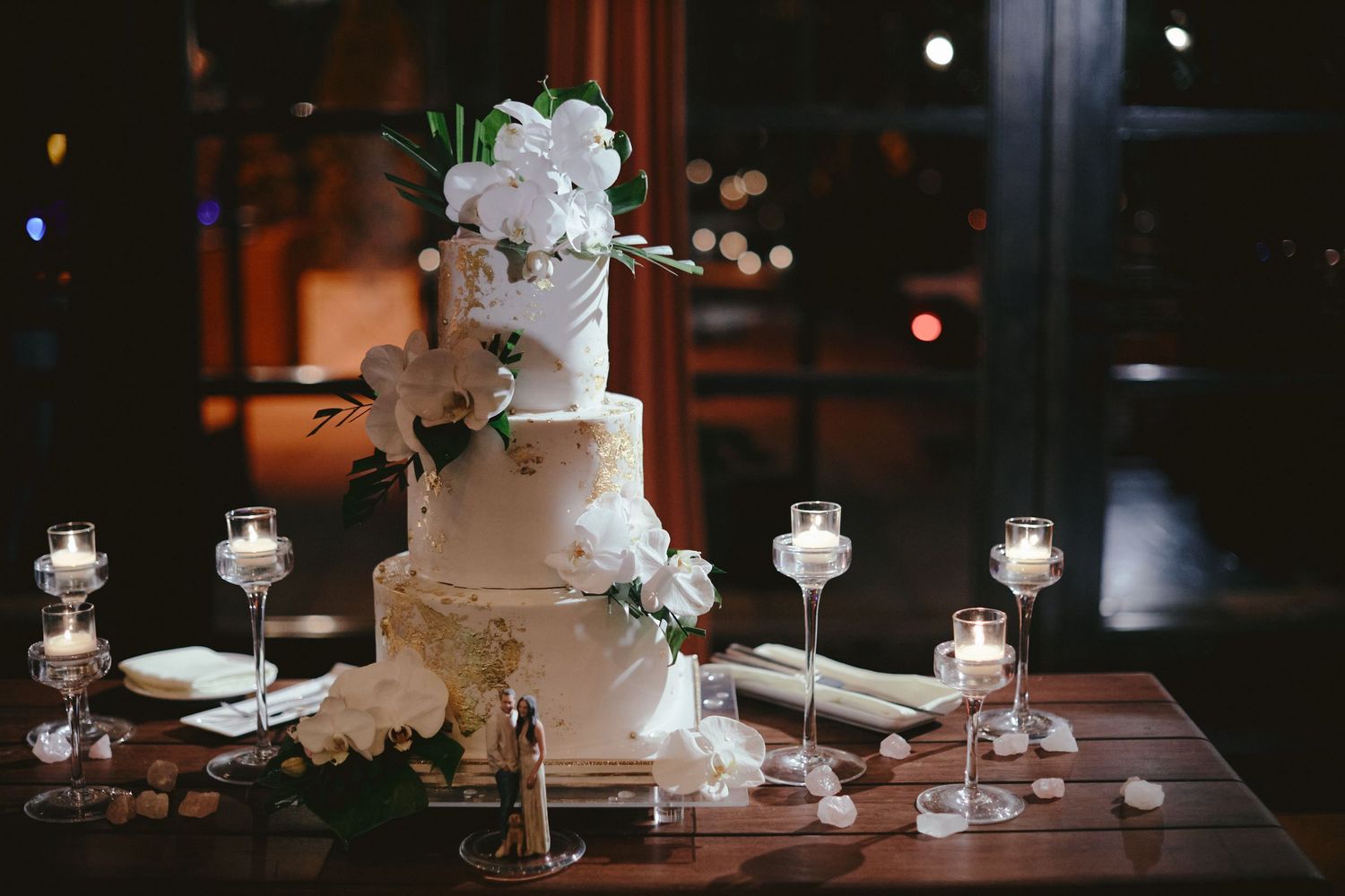 A three-tiered white wedding cake with floral decorations and glowing candles on a dark elegant table.