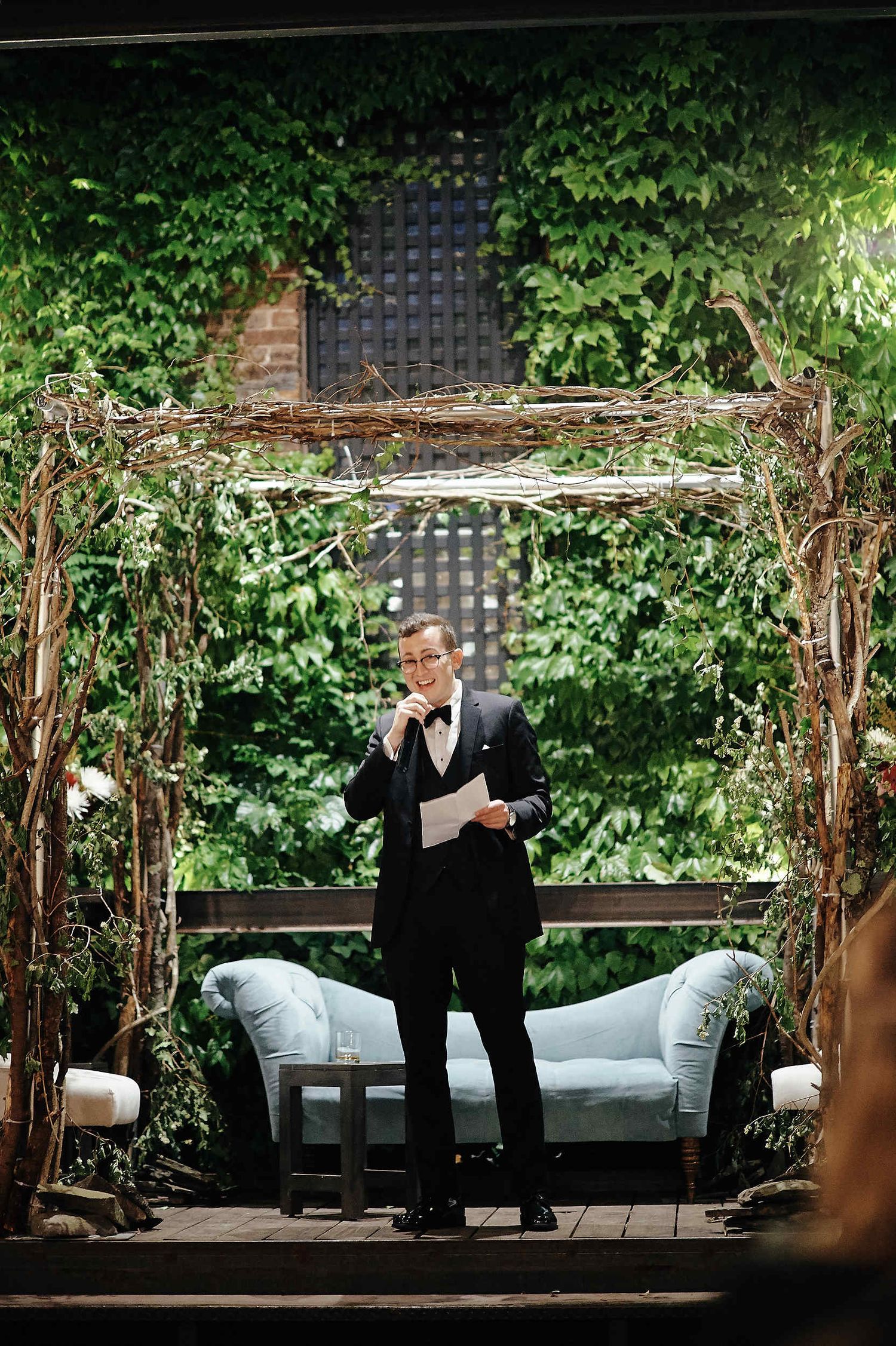 Rustic garden wedding scene with twinkle lights, greenery-covered archway and elegant white lounge seating.