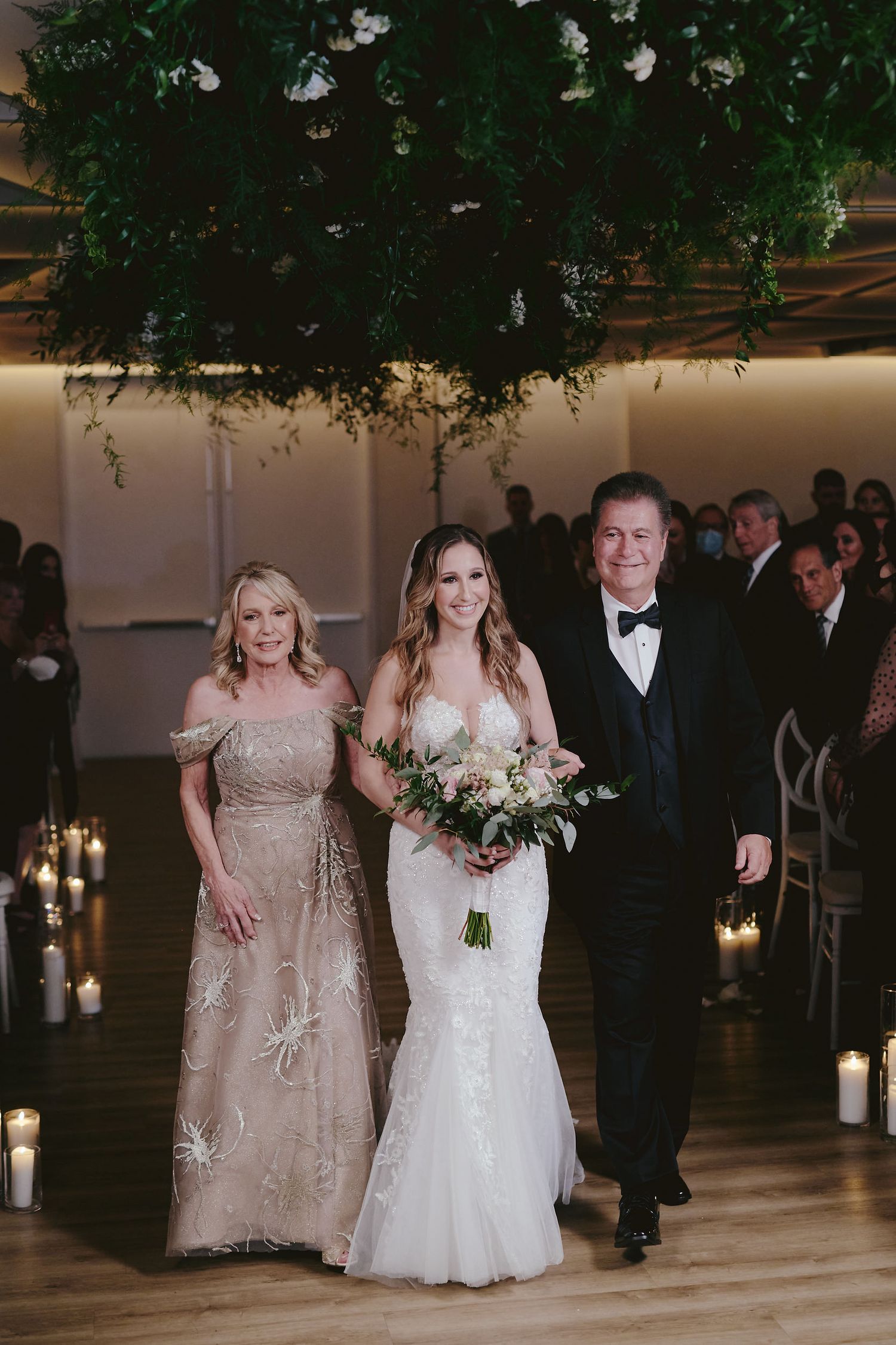 Wedding ceremony in elegant indoor reception venue with candlelit aisle and lush greenery hanging from ceiling.