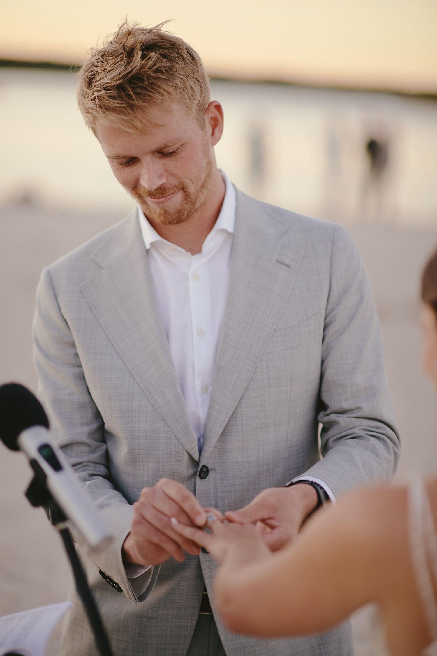 A groom in a light gray suit and white shirt looks down during an outdoor wedding ceremony.