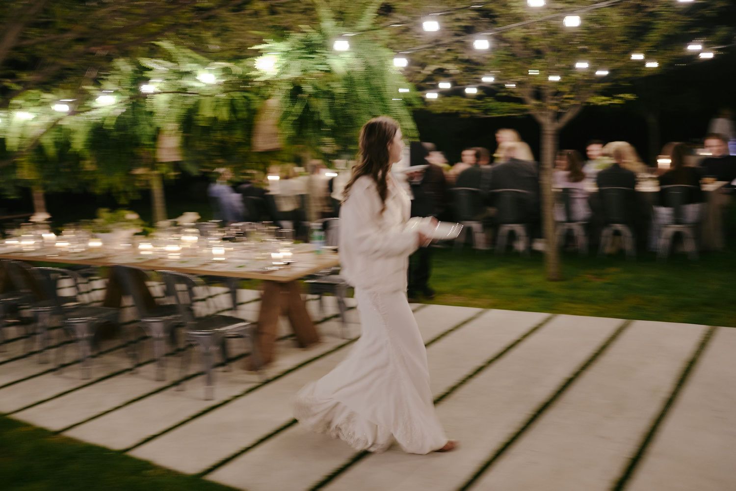 A figure in white twirls on a wooden deck under twinkling string lights during an outdoor evening celebration.